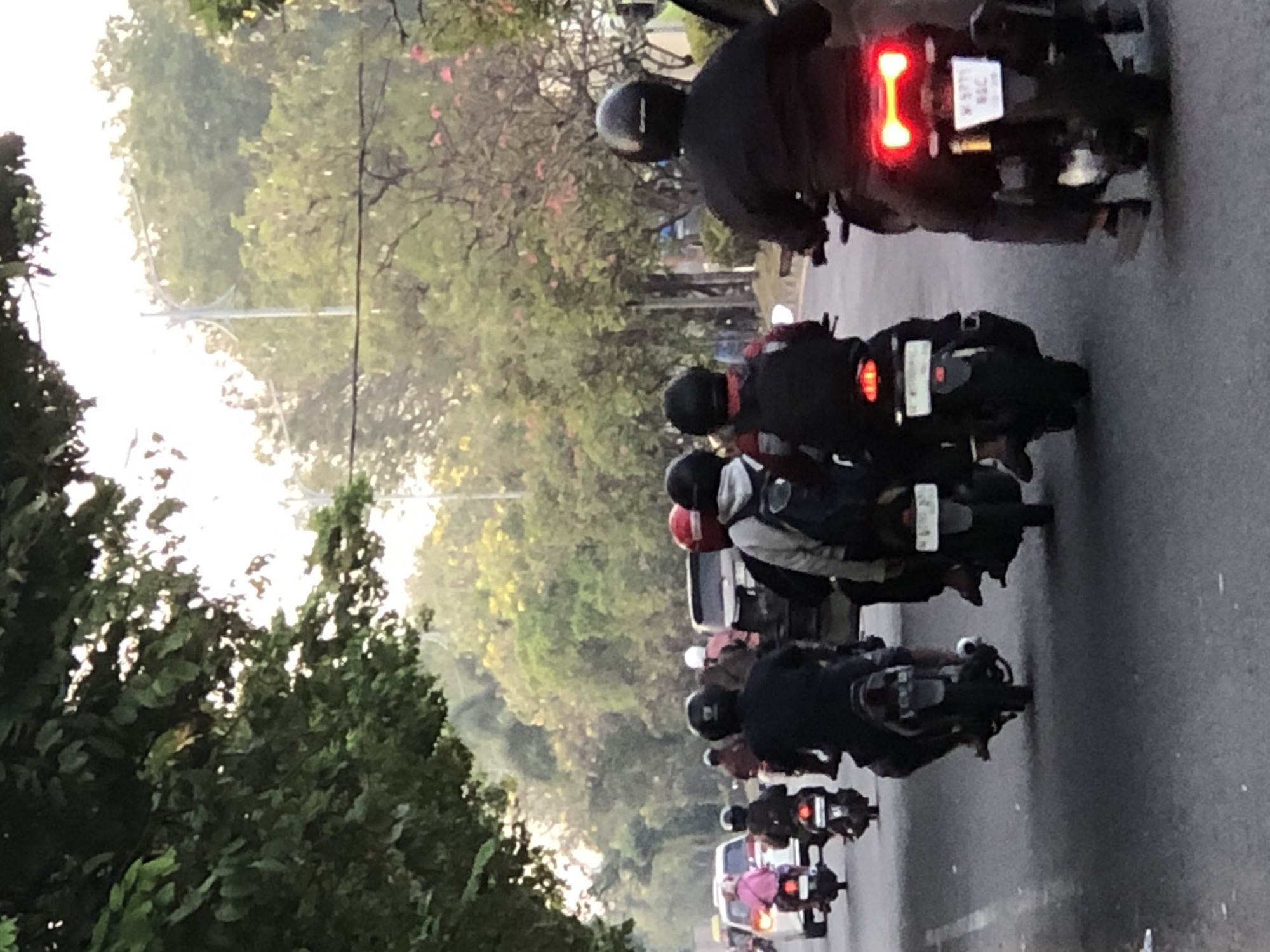 The image depicts a street scene in Ngampelsari, Indonesia, likely during daylight hours, possibly late afternoon given the soft light and shadow play. Several individuals are riding motorcycles in a traffic lane. The foreground features a motorcycle with its brake light illuminated, suggesting deceleration or presence of a rider. Behind this, other motorcycles and their riders are visible, some with helmets of various colors including black and red. In the background, there are cars and a bus, indicating a busy urban environment. The sides of the road are lined with lush green trees, some bearing pinkish-red flowers, and other foliage, suggesting a well-treed area within the city. The sky is overcast or hazy, contributing to the diffused lighting. The overall impression is of a typical commute or movement through an Indonesian city.