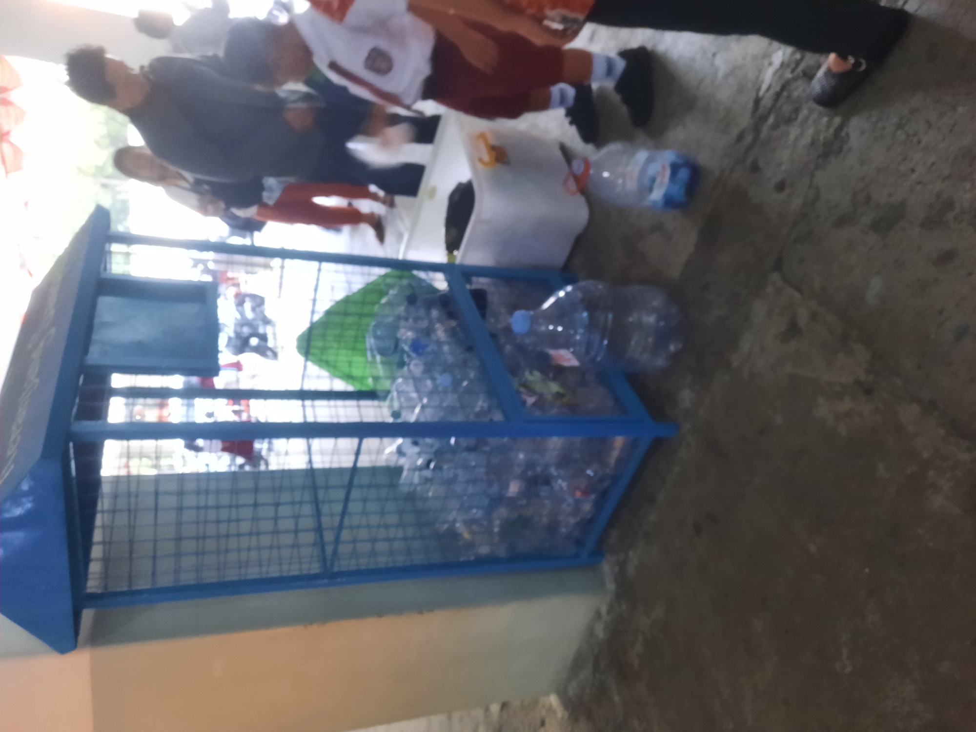 An outdoor scene in the city of North Jakarta, Indonesia, depicts a blue wire cage recycling bin filled with clear plastic water bottles. Adjacent to the bin is a white plastic tub, also containing some dark-colored items. Several people are present in the background, some of whom appear to be students in uniforms. One individual in the foreground is wearing a white shirt with a red tie and dark shorts, and another person's legs are visible in dark pants and patterned slippers. The ground is a concrete surface. The lighting suggests it is daytime.