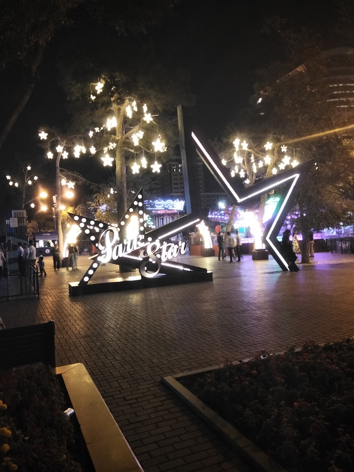 The image depicts an outdoor scene at night, likely in a public urban space such as a park or pedestrian area, with elements suggesting it is in Baku, Azerbaijan. A prominent, large, illuminated star-shaped sign with the words "Park Star" written on it is centrally located. The sign is outlined with bright white lights and dotted with smaller lights. Behind this sign, trees are decorated with numerous star-shaped lights, creating a festive and magical atmosphere. Several people are visible in the background, walking and standing, some appearing to be interacting. A young child is also present, walking towards the left side of the frame. The ground is paved with brick-like patterns. In the far background, there are buildings and more distant lights, indicating the presence of a city. The overall impression is of a well-lit, inviting nighttime environment, possibly a recreational or entertainment area within Baku.