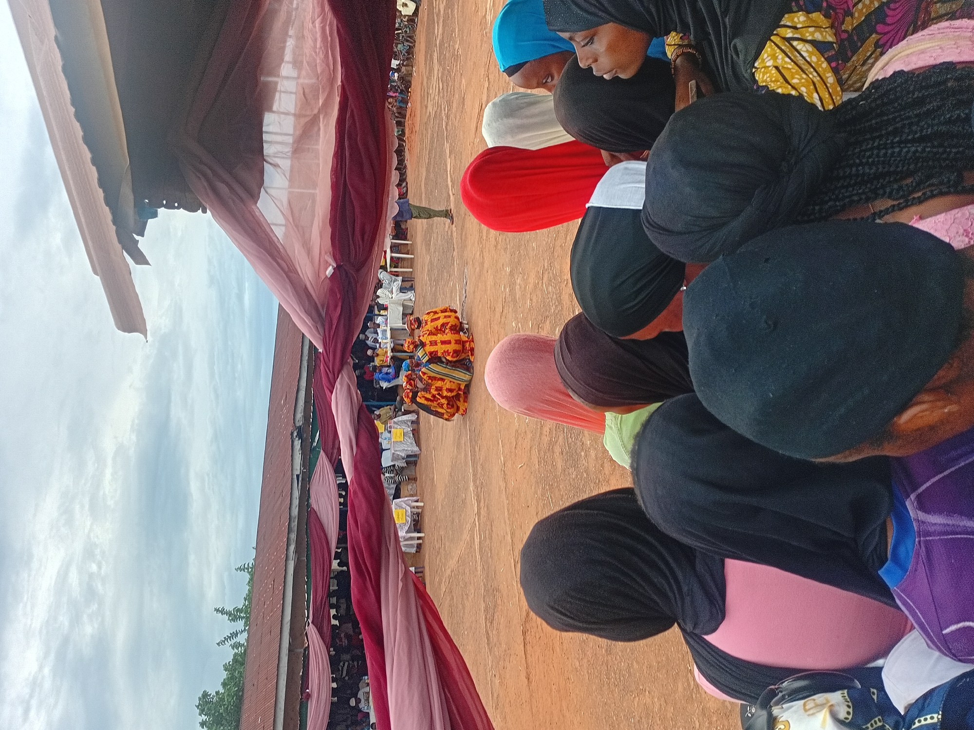 This image depicts an outdoor event or gathering in Ogbogbo, Nigeria. The scene is observed from behind a group of spectators in the foreground, looking towards a central activity area.
In the immediate foreground, the backs and profiles of approximately ten individuals, predominantly females, are visible. Most wear head coverings in various colors including black, pink, red, light blue, white, and a lighter green. Specific garment details include a pink shirt, a purple and blue striped shirt, and a yellow, orange, and pink patterned fabric on the lower right. One individual's braided hair is visible beneath a dark head covering.
The midground features an open, reddish-brown earthen ground area. Centered within this area, approximately four to five individuals, appearing to be children or young adolescents, are positioned in brightly colored, elaborately patterned costumes, predominantly yellow, orange, and red with black accents. To their right, a single adult male stands, dressed in a light blue shirt and green trousers. Along the sides of this central area, several white rectangular tables with yellow tablecloths are arranged, accompanied by chairs. Numerous individuals are seated at these tables, observing the central activity.
Spanning the upper midground and separating the foreground spectators from the background, lengths of sheer fabric in maroon and lighter pink are draped horizontally, creating an overhead canopy or decorative element. These fabrics are loosely hung and show some billow.
In the background, a long, single-story building with a reddish-brown, rusted corrugated metal roof and white eaves extends across the frame. Green foliage and trees are visible above and to the left of this structure. The sky is overcast with diffuse cloud cover.