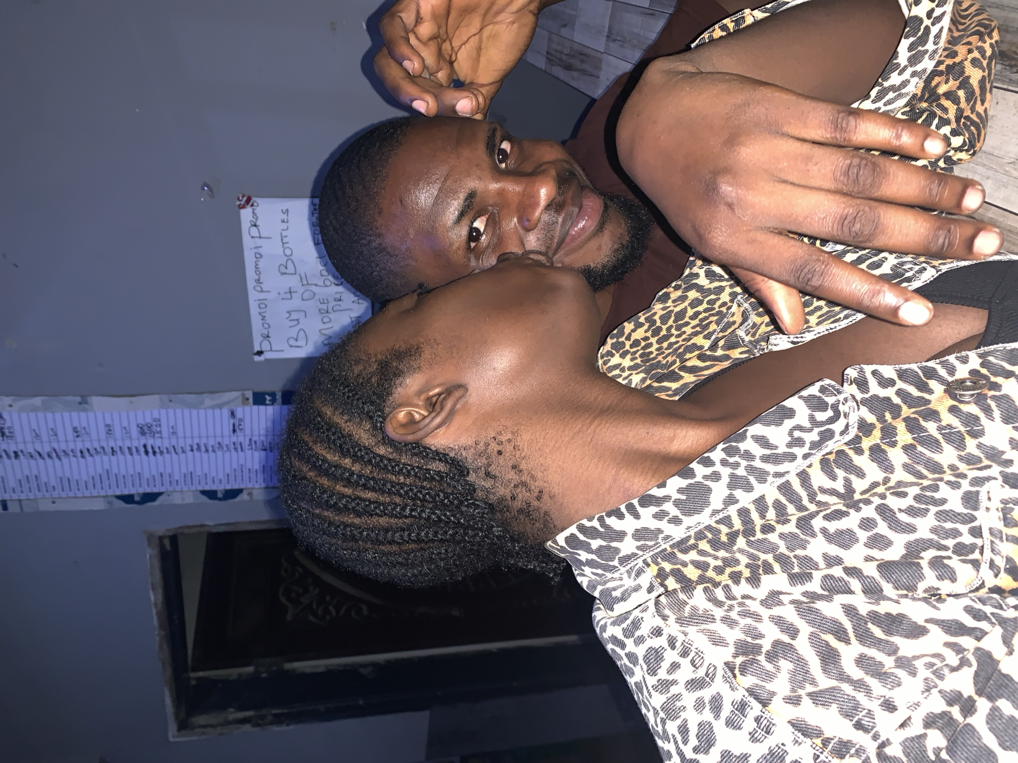 A close-up shot shows two people intimately positioned, likely within an indoor setting in New Stadium, Nigeria. The lighting appears to be ambient and possibly fluorescent. One individual, a man with a dark beard and short-cropped hair, gazes toward the camera with a slight smile. The other person is angled to the side, seemingly about to kiss the man's face. Both individuals are wearing garments with a leopard print pattern. Above them, a handwritten note with the words "Promo Promo Promo - Buy 4 Bottles of MORE Bool For The Price" is affixed to the wall, indicating a promotional deal. A small, framed object hangs nearby, partially obscured.