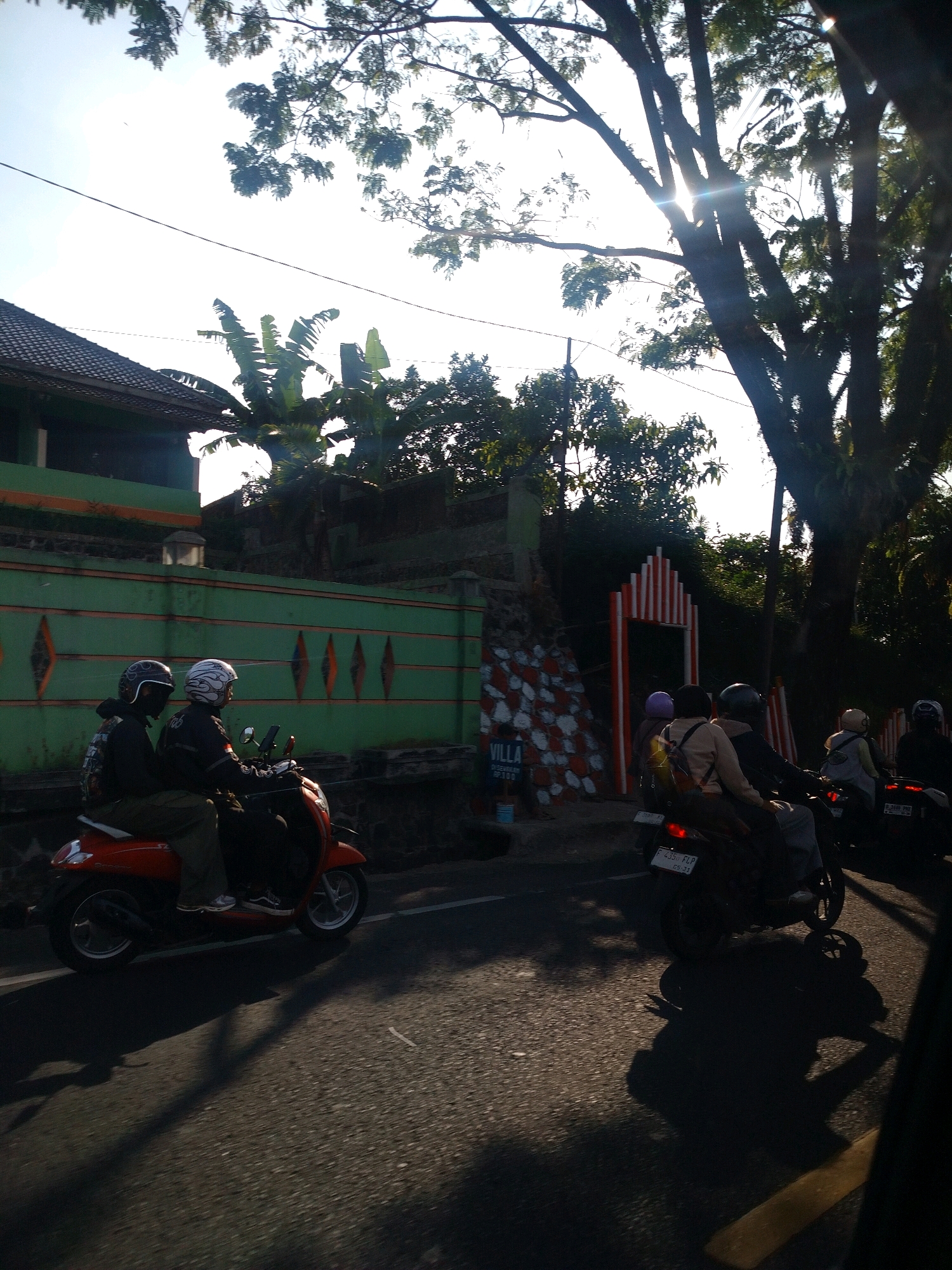 Two individuals wearing helmets are riding a red scooter down a paved road. The person in the back is wearing a dark jacket and gray pants, while the person in the front is wearing a black jacket and black pants. To the left of the scooter, a green wall with diamond-shaped cutouts runs alongside the road. A building with a tiled roof is visible behind the wall. To the right of the scooter, there is a decorative archway with red and white stripes, and a stone wall with red and white patterns. The road is illuminated by sunlight filtering through the trees. Further down the road, other individuals on scooters are visible. The environment appears to be a semi-urban area with lush vegetation, suggesting a location like Cisarua, Indonesia, known for its scenic beauty and natural attractions.