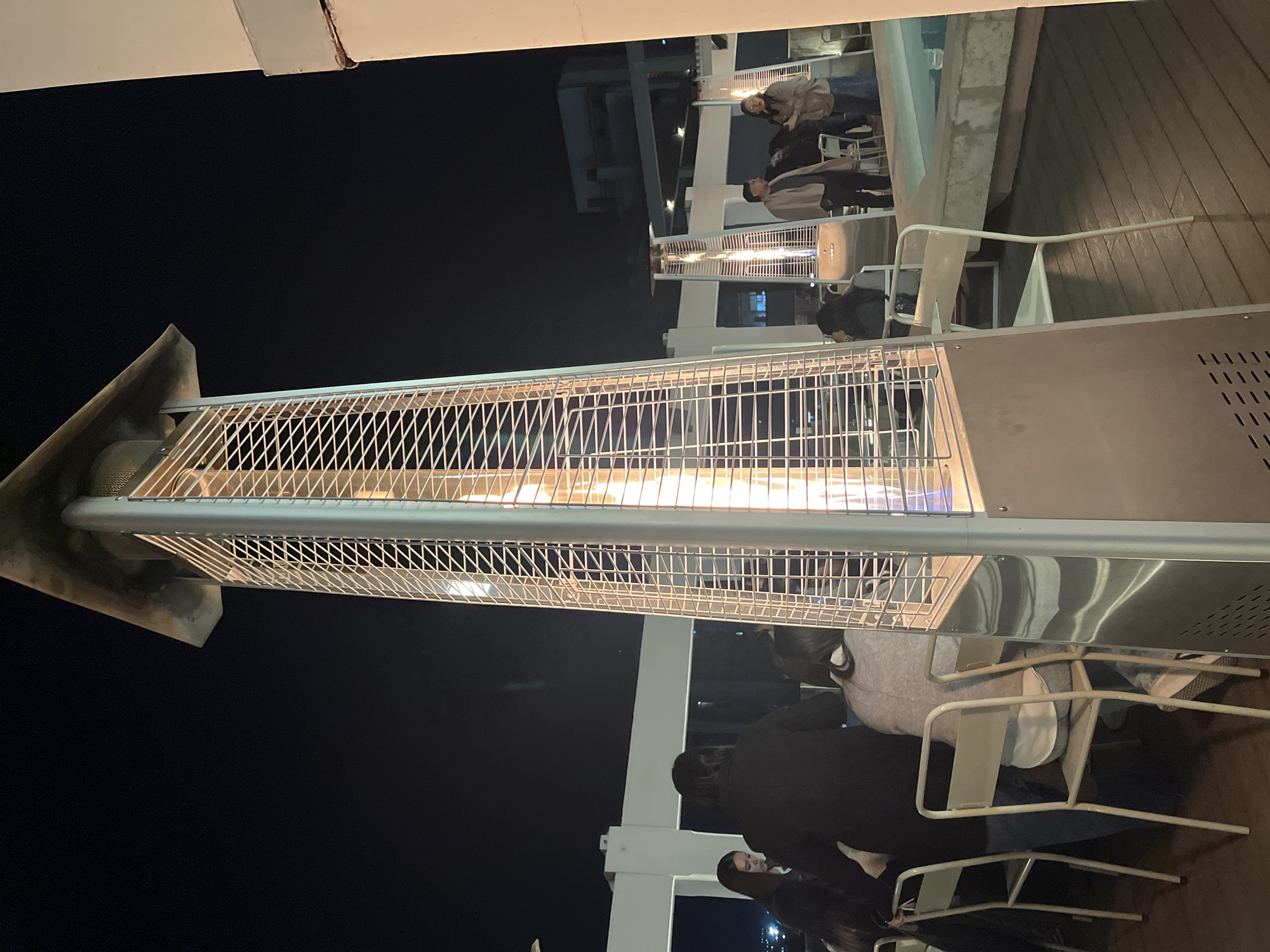 The image depicts an outdoor setting in San Fernando del Valle de Catamarca, Argentina, at night. The scene is illuminated by a row of standing patio heaters, with visible flames inside their protective cages. A group of people are seated at tables near the heaters, presumably dining or socializing. The background features a dimly lit building, suggesting a commercial or residential area. The immediate foreground consists of the patio area, including chairs and table settings.