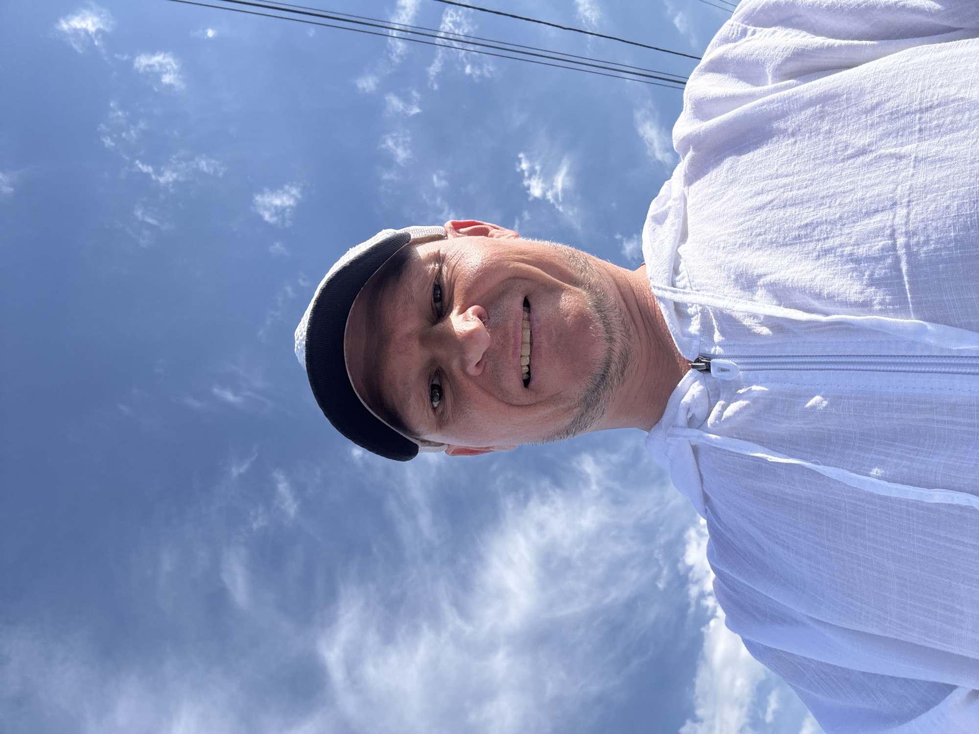 In Zharbulak, Kazakhstan, under a bright blue sky dotted with wispy clouds, a man wearing a white hooded jacket and a baseball cap is captured in a low-angle, close-up selfie. He has a light complexion and is smiling, revealing his teeth. The background shows utility lines stretching across the sky, typical of an urban or semi-urban setting. The man's jacket is zipped up, and its textured fabric is visible, contrasting with the smooth sky.