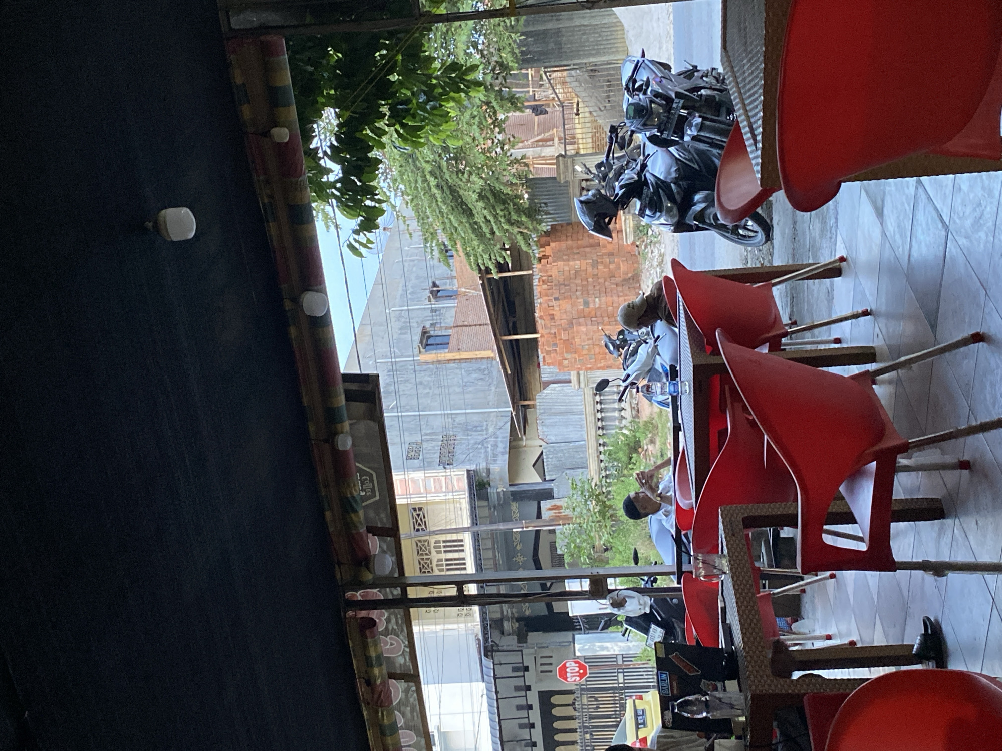 This is an outdoor shot of a cafe in Lhokseumawe, Indonesia, taken during the daytime. The foreground is dominated by several red plastic chairs and tables, suggesting a casual seating area. In the mid-ground, two people are seated at a table, one appearing to be on their phone. Behind them, several motorcycles are parked, indicating this might be a popular spot for riders. Beyond the cafe, a street scene unfolds with buildings, a STOP sign, and piles of bricks, possibly from ongoing construction. Lush green trees provide a natural element to the urban landscape. The overall impression is of a functional, community-oriented space in a bustling Indonesian city.