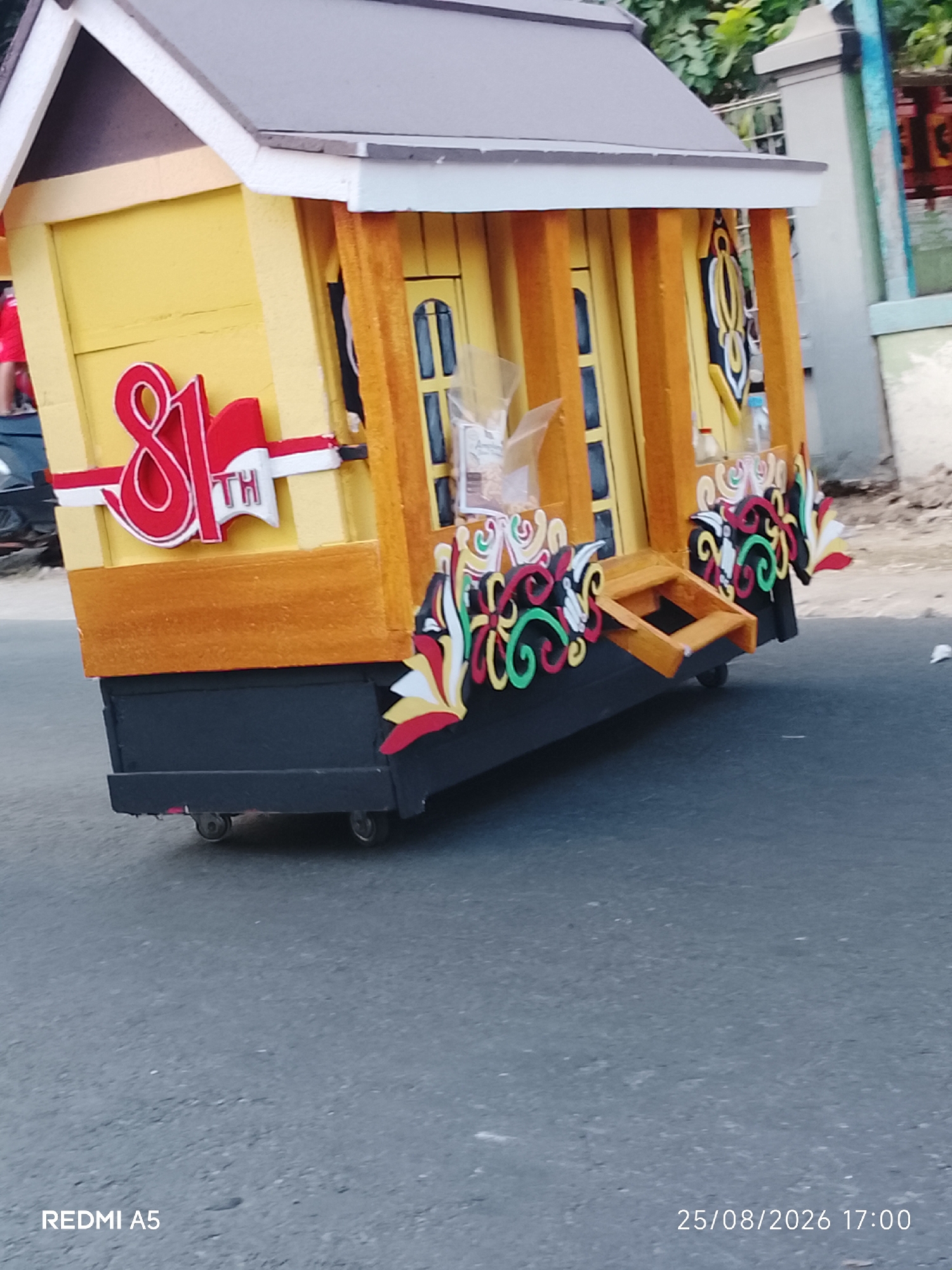 A yellow miniature house on wheels is moving on a paved road in Semanding, Indonesia. The house has a dark gray roof and a dark gray base with small wheels. It is decorated with red and white flags, as well as colorful ornamental patterns on the sides. The time stamp "25/08/2026 17:00" and "REDMI A5" are visible at the bottom of the image. The background shows some green foliage and a light-colored wall.