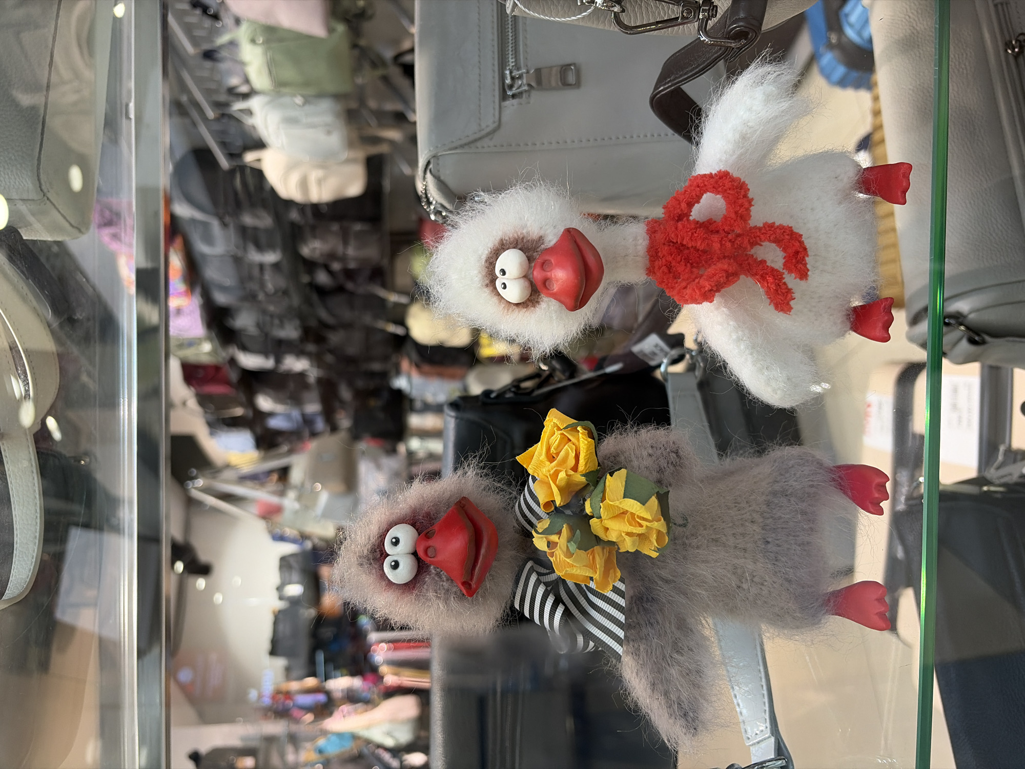 The media file, captured in Izhevsk, Russia, displays two fluffy, bird-like plush toys presented horizontally within a glass display, likely a shop window or case. Both toys exhibit exaggerated, comical features, including large, round white eyes with small black pupils and prominent, glossy red beaks or mouths.
The upper toy is predominantly white with a shaggy, soft texture, adorned with a fluffy red bow on its chest. Its two small, red, webbed feet are visible.
Positioned beneath it, the second toy is greyish-brown and also features a shaggy texture. It is decorated with a black and white striped ribbon tied into a bow, from which a cluster of small, yellow paper roses emerges. This toy also has two red webbed feet.
The background behind the display is blurred, revealing shelves and racks holding numerous handbags and other retail items, suggesting a setting within a shop, boutique, or market. Reflections on the glass obscure some background details. The time of day appears to be daytime. There is no visible text in the image.