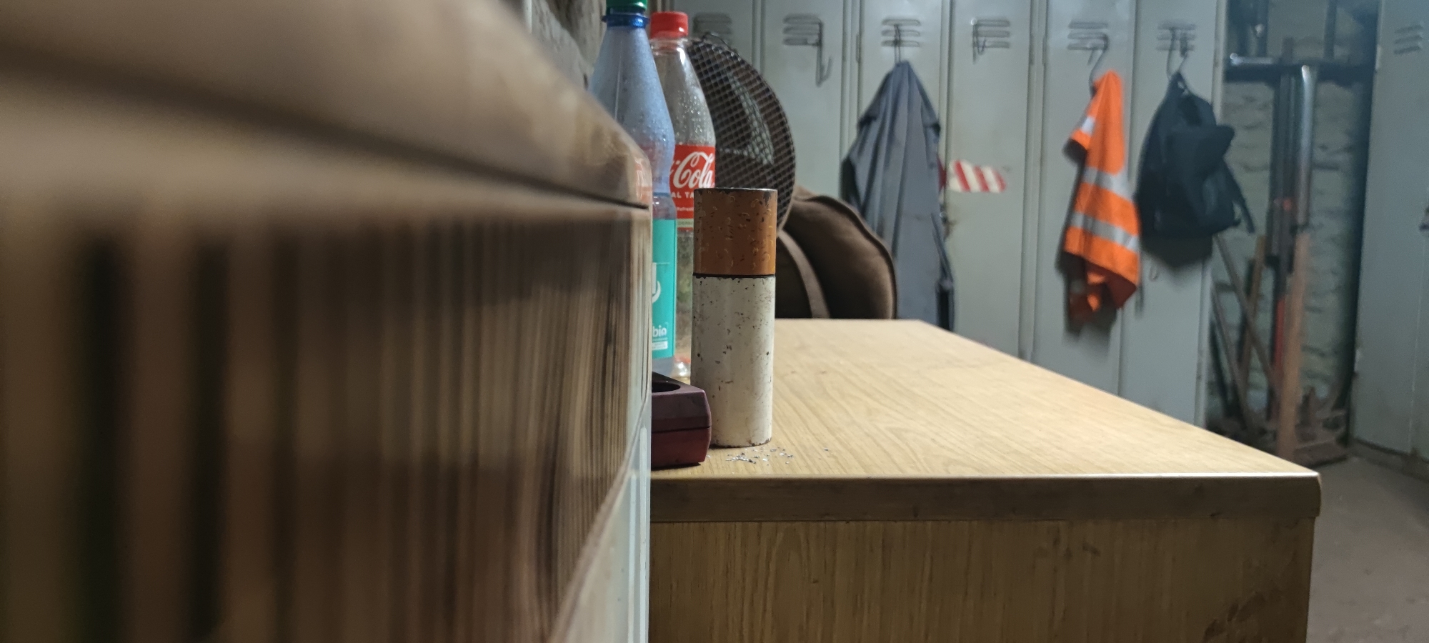 The image shows a close-up, low-angle view of a wooden desk with several items on it. To the left, a portion of a wooden cabinet or drawer is visible, its surface showing vertical wood grain. On the desk, in the center of the frame, is a cylindrical object, possibly a cigarette butt or a small container, with a brown top and a speckled white body. Behind it, two plastic bottles are lined up: one is a blue bottle with a green cap, and the other is a clear bottle with a red cap, showing the Coca-Cola logo. To the right of the bottles, a dark brown leather bag is partially visible. In the background, a row of metal lockers is lined up against a wall. Several items are hanging on or near the lockers: a grey jacket hangs on a hook, an orange high-visibility vest with reflective stripes is on another hook, and a black backpack is also hung up. The environment appears to be a locker room or a changing area, possibly in an industrial setting or a sports facility, located in Salzgitter, Germany. The lighting is dim, creating shadows and highlighting the textures of the objects on the desk.