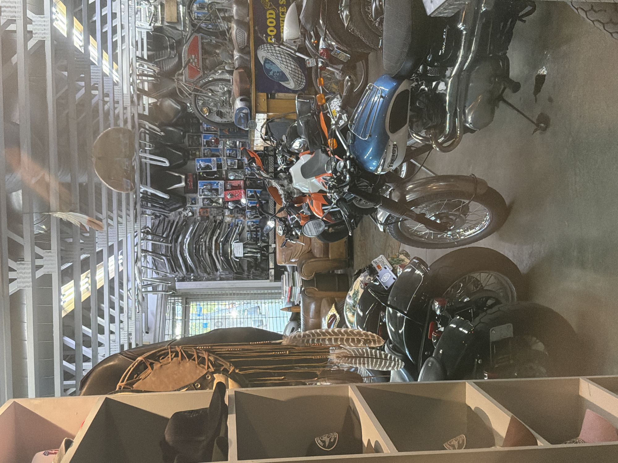 The image shows the interior of a cluttered space, potentially a shop or garage, filled with motorcycles and motorcycle parts. The location is West Hollywood, United States of America. Various motorcycles of different models and ages are parked and stored throughout the area. Motorcycle parts such as handlebars, wheels, and accessories are organized and displayed. The background includes shelves containing motorcycle-related merchandise, such as hats with logos. The overall environment suggests a motorcycle dealership or storage facility.