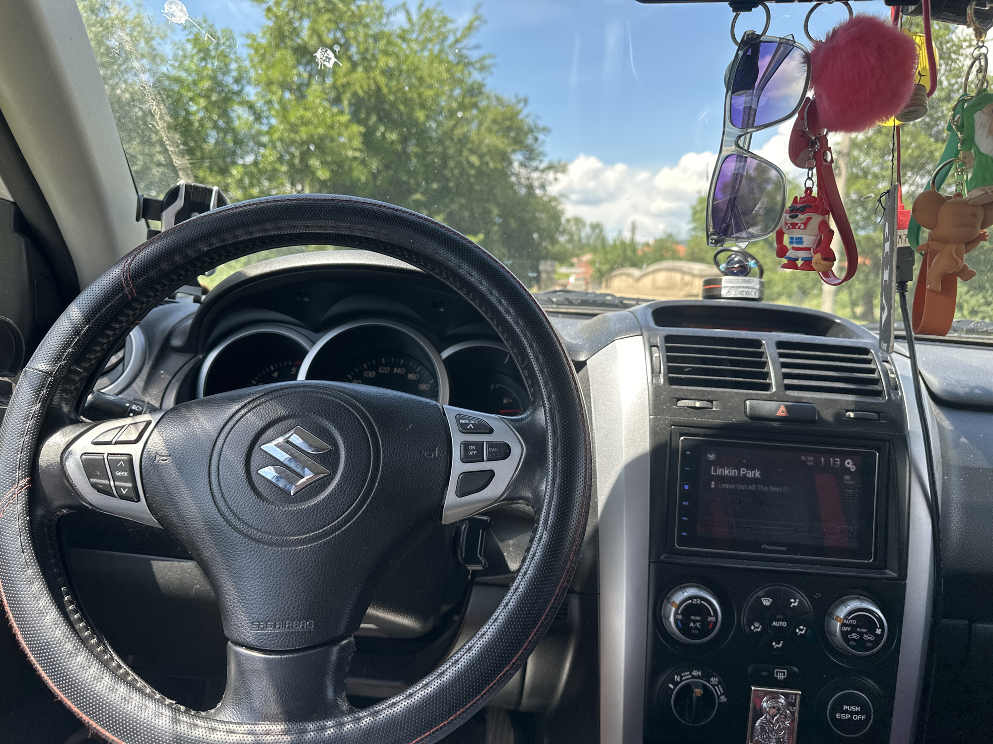 The image shows the interior of a car, likely a Suzuki Grand Vitara, from the perspective of the driver's seat. A black steering wheel with a Suzuki logo in the center is prominent on the left. To the right of the steering wheel is the car's dashboard, featuring an infotainment screen displaying "Linkin Park" and "Leave Out All The Rest". Below the screen are various climate control knobs and buttons, including a dial for A/C temperature and fan speed, and another for the automatic climate control system. A gear shifter is visible further down. Several items are hanging from the rearview mirror, including a pair of sunglasses, a pink fluffy pom-pom, and several novelty keychains depicting characters such as Taz from Looney Tunes and Jerry from Tom and Jerry. Outside the car, through the windshield, trees and a blue sky with scattered clouds are visible. The overall impression is of a well-used vehicle, possibly parked or driving on a sunny day. The metadata indicates the image was taken in Cherven Breg, Bulgaria.