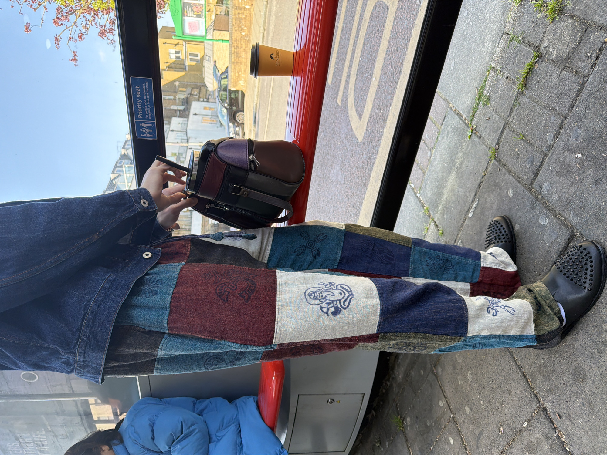 This image shows a person on a bus in London, United Kingdom. The person is wearing a blue denim jacket and patchwork pants in various colors, with black shoes with a studded design. They are holding a black object, possibly a camera, with a brown and purple bag resting on the bus ledge next to a coffee cup. The bus interior has a red safety bar and a “priority seat” sign on the window. Outside the window, buildings are visible on a sunny day, and there are typical London street signs and vegetation. The bus is stopped on a paved street with a few weeds growing in the cracks. Another person is visible wearing a blue coat.