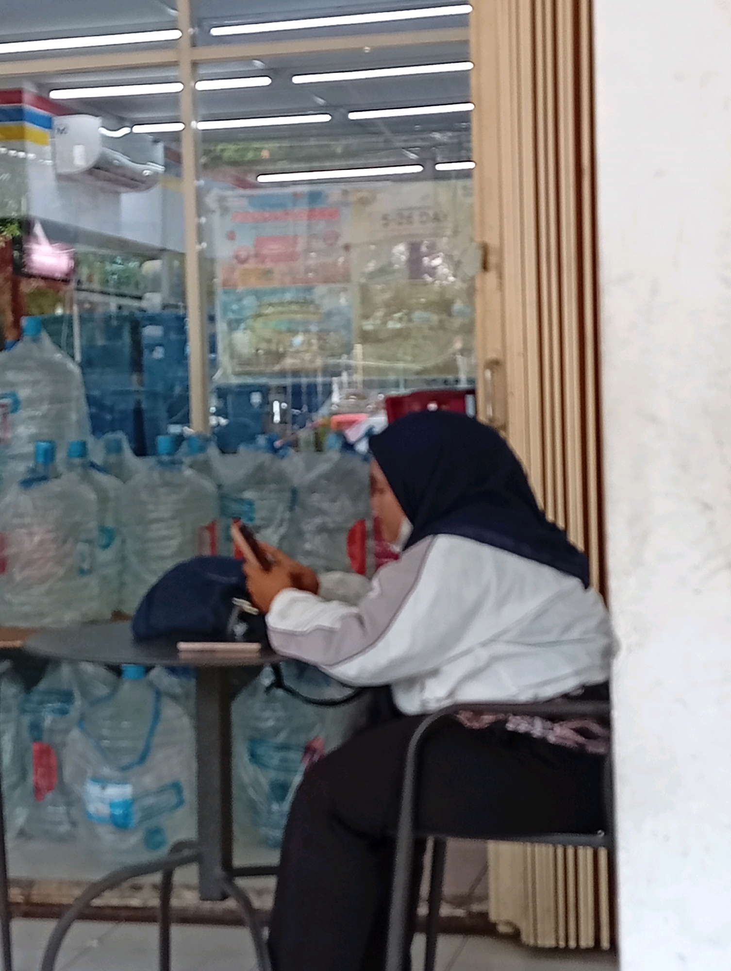 The image depicts an outdoor scene, likely in front of a shop in Surabaya, Indonesia. A young woman wearing a dark blue hijab and a light-colored jacket is seated at a small, round table, looking at her phone. She is dressed in dark pants. On the table, there's a dark blue bag and a cell phone. Behind the table, numerous large plastic bottles, presumably containing water, are stacked. The background reveals the interior of a shop with shelves and displays visible through the glass window. The shop has a light-colored metal grate door. The lighting suggests it is daytime.