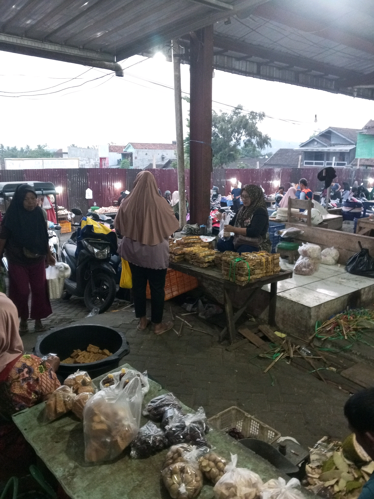 The image depicts an outdoor market scene in Gentan, Indonesia, likely a traditional market setting. In the foreground, various food items are displayed on a green tarped table. These include packaged snacks or dried goods in plastic bags, some appearing to be fried tempeh or crackers, and a large black tub filled with similar food items. To the left, a woman wearing a dark headscarf and maroon pants is walking away from the camera, holding a white plastic bag.
Behind the table, a scooter is parked. Further back, two women are engaged in transaction or conversation. One woman, facing away from the camera, is wearing a light brown headscarf and dark pants, holding a yellow plastic bag. The other woman, facing the camera, is wearing a patterned headscarf and dark clothing, seated behind a wooden table displaying stacks of packaged food items, possibly snacks or dried goods, bound with green string.
The market appears to be covered by a corrugated metal roof, supported by wooden beams and poles. Beyond the market stalls, a fence made of corrugated metal and wood separates the market from a residential area. Several buildings with pitched roofs are visible in the background, along with trees and utility poles. The lighting suggests it is either early morning or late afternoon. The overall atmosphere is that of a bustling local market.
