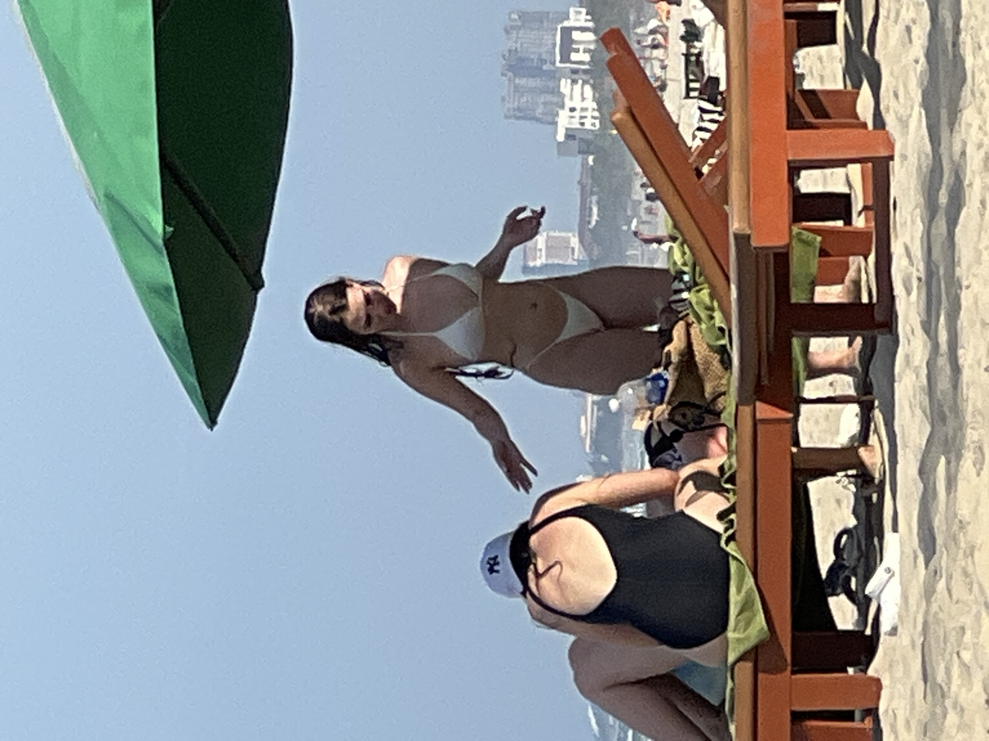 The image captures a beach scene in Da Nang, Vietnam, on a sunny day with a clear blue sky. In the foreground, two individuals are seated on orange lounge chairs. One person, on the left, wears a black swimsuit and a white baseball cap with a black logo. The other individual, partially visible, is seated next to them.
Standing on the sand between the lounge chairs is a person in a white bikini, facing the viewer's right. The person is reaching out with their right hand, their fingers splayed. Behind them, the cityscape of Da Nang is visible in the distance, with modern buildings and structures lining the coast. A large green umbrella is positioned in the upper right corner of the frame, casting a shadow. The beach is sandy, and other beachgoers and lounge chairs are scattered further in the background.