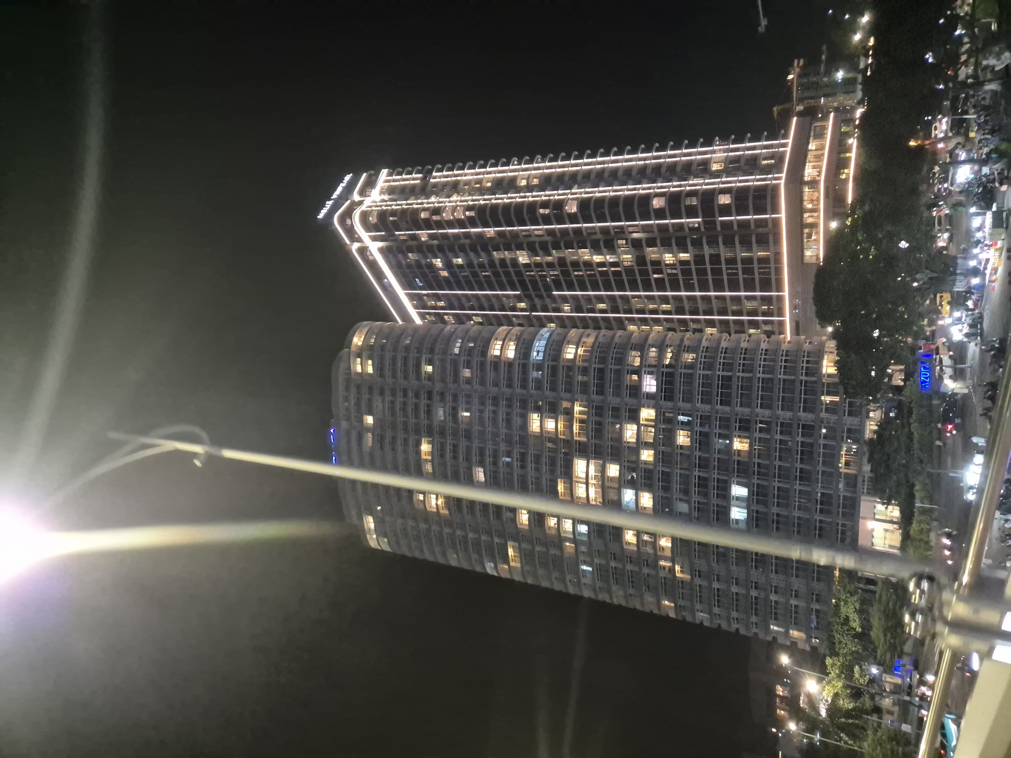 The image captures a night scene in Da Nang, Vietnam, featuring the Melia Vinpearl building, a tall, modern skyscraper illuminated by white lights along its edges and horizontal bands. Adjacent to it is another similarly designed skyscraper, also with lights visible in its windows. The buildings are set against a dark night sky. In the foreground, a street or promenade is visible with trees, streetlights, and a busy gathering of people. Several vehicles, including cars and motorbikes, are present, with their headlights and taillights creating streaks of light. The overall impression is of a vibrant urban environment at night.