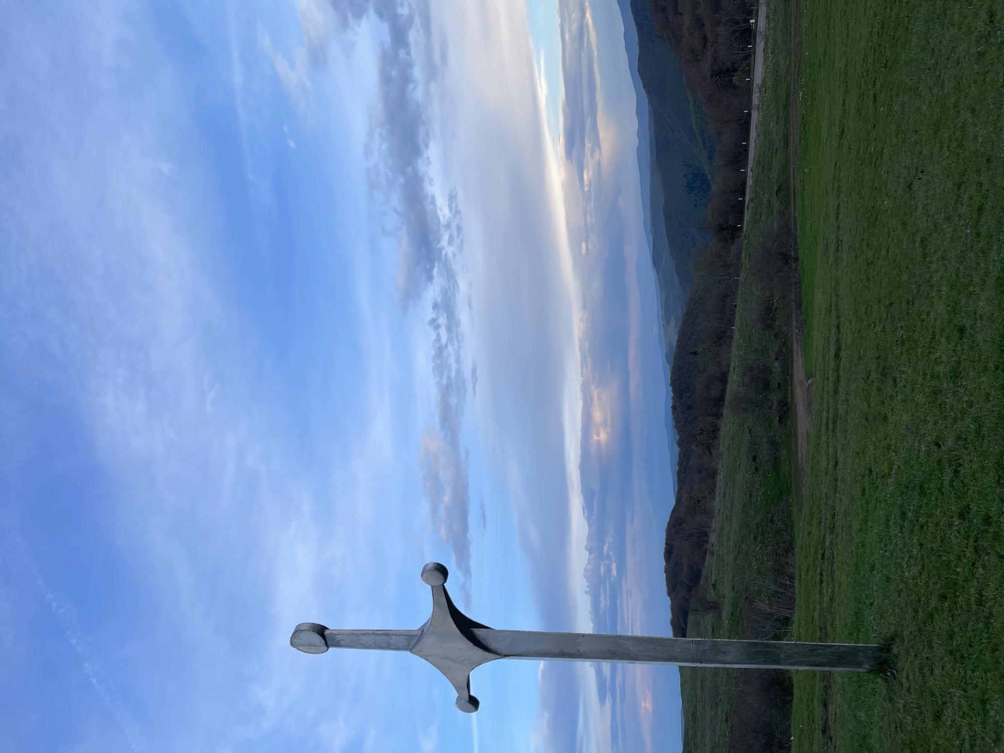 A long sword monument stands upright on a grassy field in Shamta, Georgia. The sword is metallic, with a rounded pommel and crossguard. The background shows rolling hills covered in trees and a blue sky with light clouds.