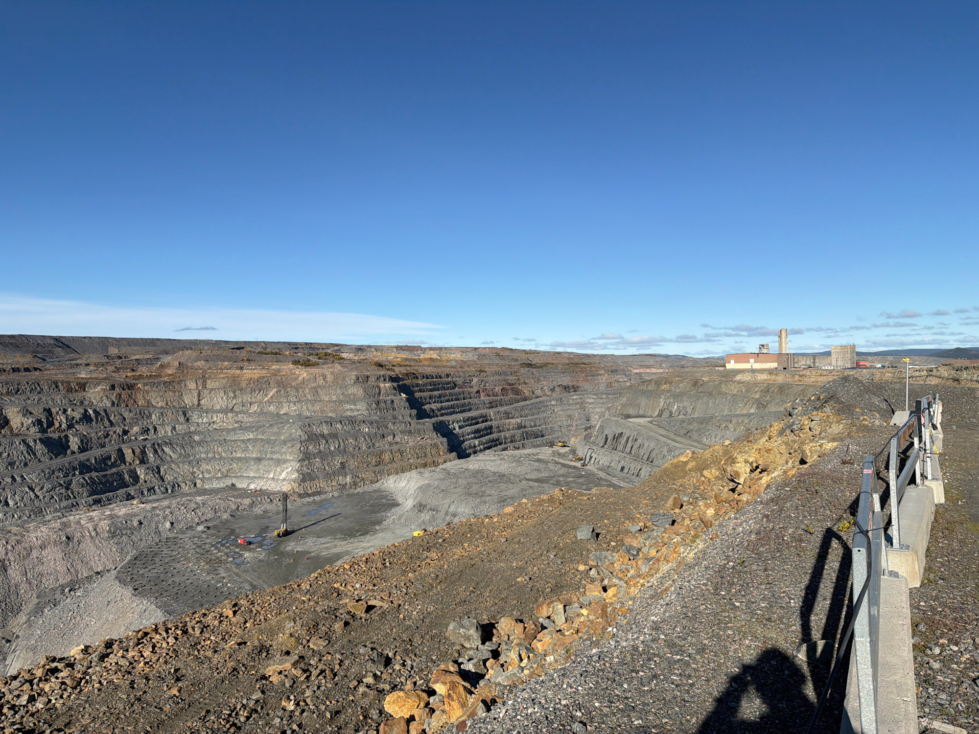 The image shows an aerial view of a large open-pit mine located in the vicinity of Gällivare, Sweden. The mine is characterized by its vast, stepped excavation, revealing layers of rock and earth. In the foreground, a gravel path runs along a metal railing, casting long shadows onto the ground. The path is bordered by piles of loose rocks and debris. In the middle ground, the mine floor is visible with evidence of ongoing operations, including what appears to be drilling equipment and a yellow excavator. The backdrop features industrial buildings with a tall smokestack, suggesting processing facilities associated with the mine. The sky is a clear, bright blue.