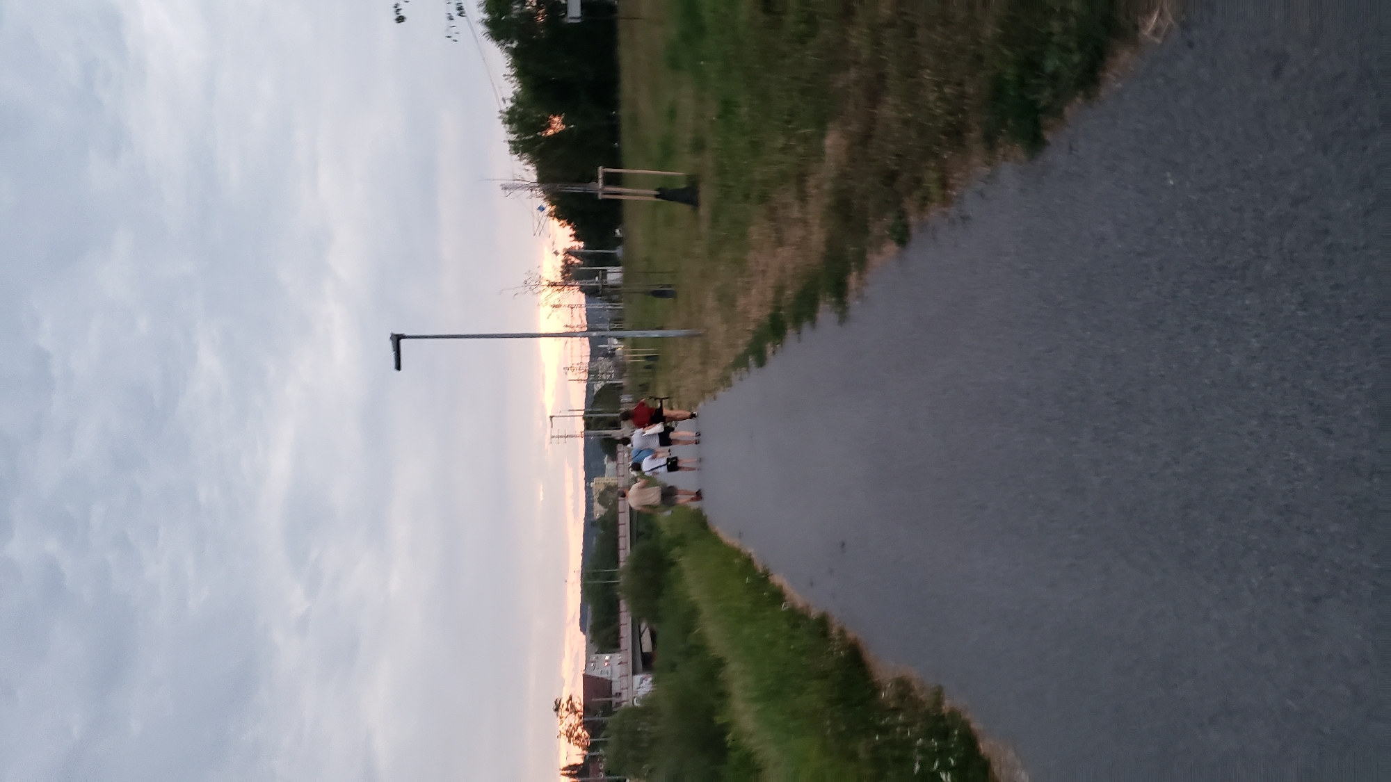 A group of four people are walking away from the camera down a paved path on a cloudy day. The path is bordered by grassy areas on both sides. In the background, there is a bridge and some buildings, suggesting an urban or suburban setting. The sky is overcast with a soft, diffused light, typical of twilight. The overall scene is peaceful and suggests a leisurely stroll. The image appears to have been taken in the city of Prešov, Slovakia.
