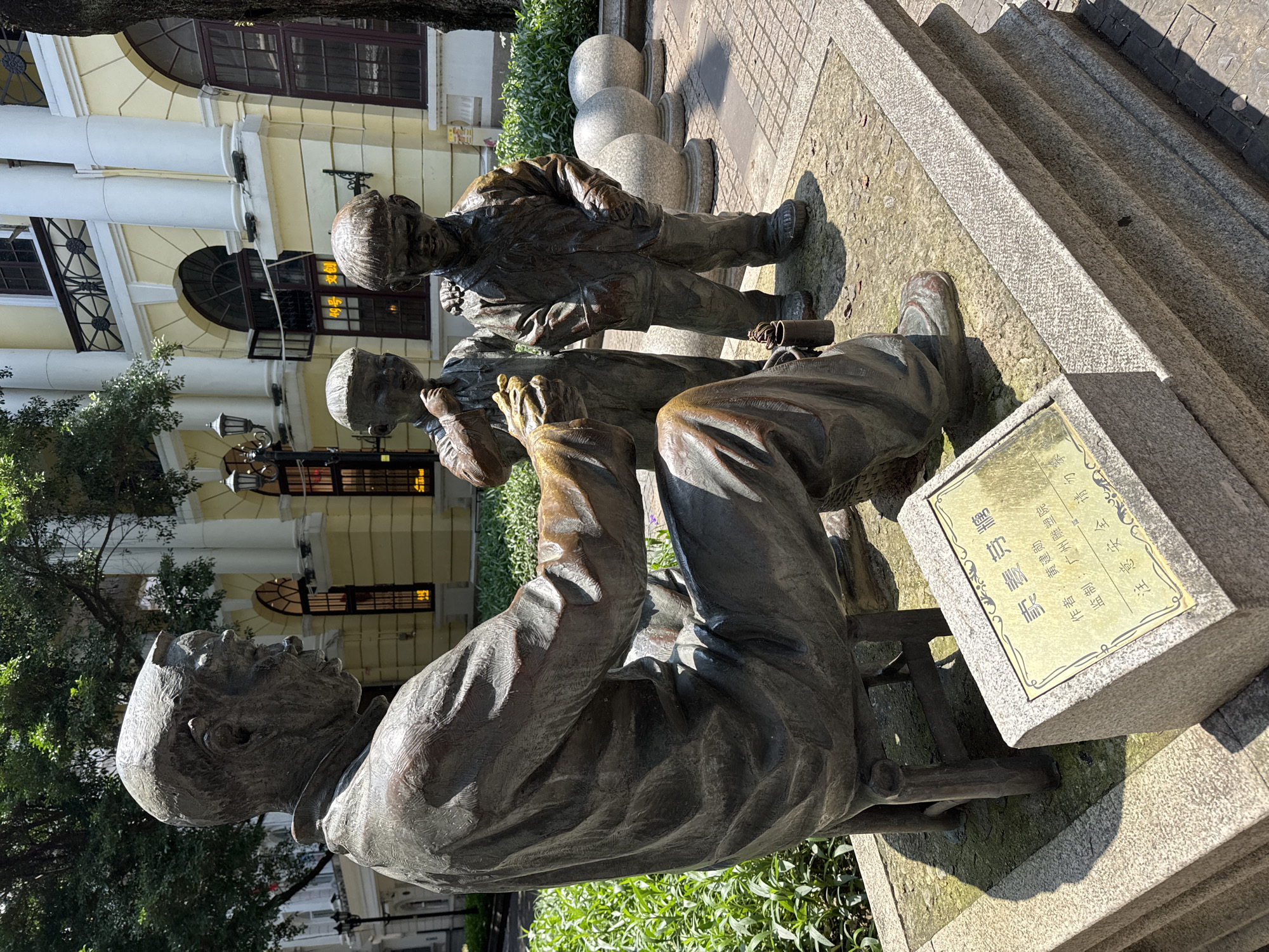 The image captures a bronze statue of a seated man with two children beside him, presumably selling malt sugar. The statue is situated outdoors on a paved area with green foliage surrounding it. Behind the statue is a classical-style building with columns and arched windows. A plaque is placed next to the statue, indicating the artist, Huang Jianxun, the production company, Guangzhou Sculpture Institute, and a warning not to climb the statue. Based on the plaque, it can be inferred that the statue is located in Guangzhou, Liwan District, China.