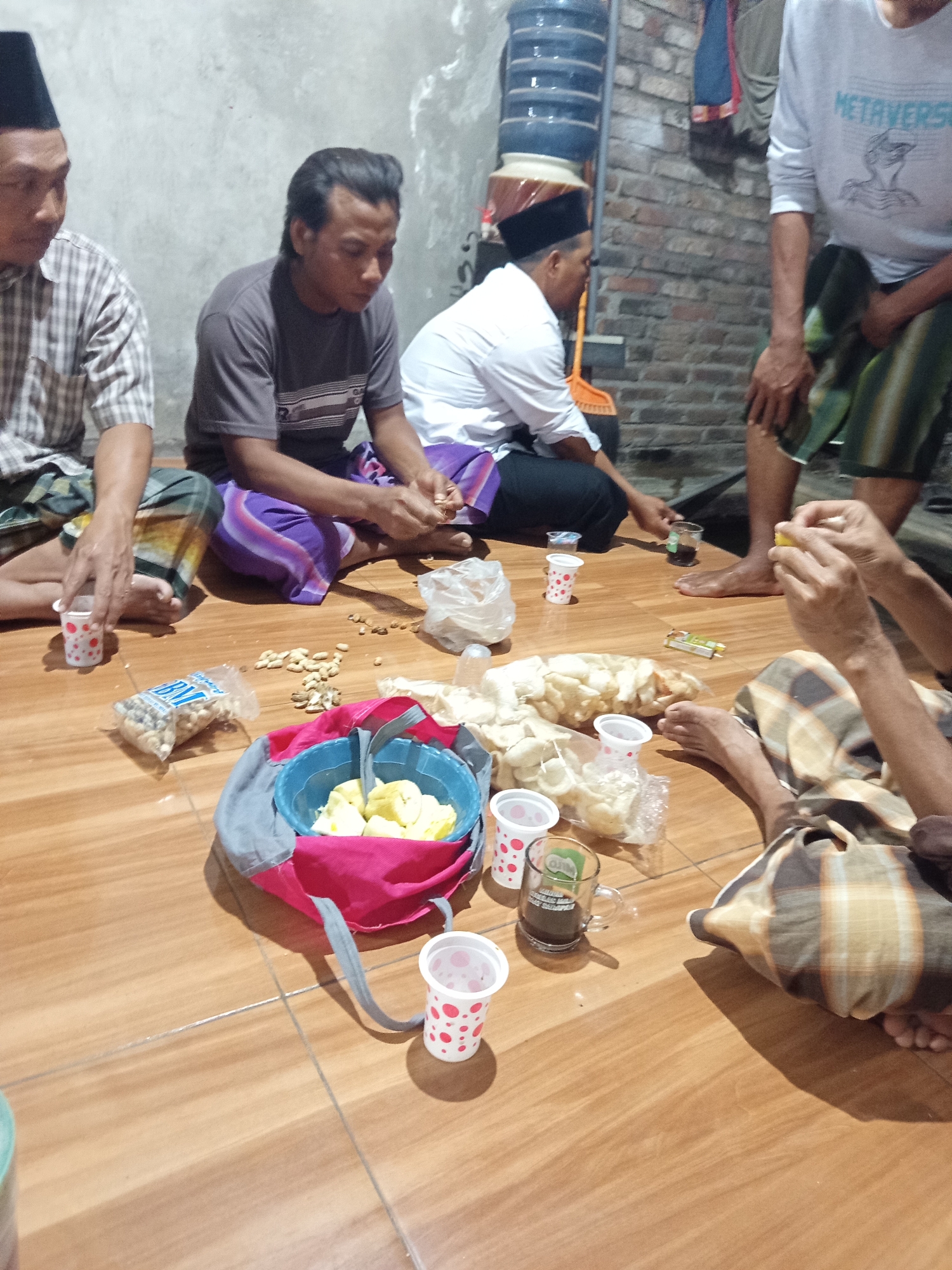 The image depicts a gathering of four men sitting on a wooden floor indoors, likely in Dermo, Indonesia. The scene appears to be informal, with food and drinks laid out on the floor.
In the foreground, to the left, a man wearing a black peci and a checkered shirt is seated, holding a small white cup with red polka dots. Next to him is a partially opened plastic bag containing small, round objects, possibly nuts or seeds.
To the right of the first man, another man in a grey t-shirt and a sarong is visible. He is looking down and appears to be interacting with some scattered nuts on the floor. In front of him, a blue bag contains yellow fruit pieces, and several small white cups with red polka dots are scattered around. There are also some packaged snacks and a glass of dark liquid.
Further back, a man in a white shirt and dark pants is seated. He is partially obscured by other individuals. To the far right, a person in a white long-sleeved shirt with the word "METAVERSE" printed on it is standing or kneeling, wearing a patterned sarong. Their legs are visible in the lower right portion of the image.
The environment is simple, with a concrete wall in the background and a large water dispenser unit to the left. The floor is made of polished wooden planks. The overall impression is of a casual social gathering or a meal shared amongst friends or family.