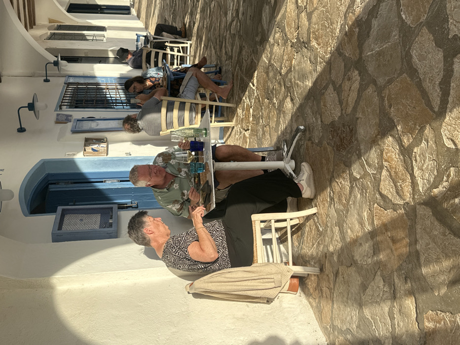 The image displays an outdoor dining area within an arched white colonnade in Palafrugell, Spain. The ground is paved with irregular, light-toned flagstones, with direct sunlight illuminating the left side and casting distinct diagonal shadows across the pavement. The architectural elements consist of white stucco walls, blue painted wooden doors, window frames with grilles, and wall-mounted blue light fixtures. A blue, patterned box-like object is mounted on the wall above the foreground blue door.
In the foreground, an older woman with short grey hair, wearing a patterned short-sleeved top and dark trousers, sits on a white wooden chair. A beige cardigan is draped over the chair's back. She holds cutlery over a plate on a white square table. Opposite her, an older man with short grey hair and glasses, wearing a patterned green short-sleeved shirt and light shorts, is also seated at the table, eating from a plate. The table holds additional items including a wine glass, a blue tumbler, and a clear glass bottle.
In the mid-ground, a second white square table is occupied by a man in a grey shirt and a woman holding a smartphone. The woman is barefoot. Further down the colonnade, a third table shows a man in a dark hat seated with his back partially to the viewer. The background features more white walls and blue accents, illuminated by direct sunlight.