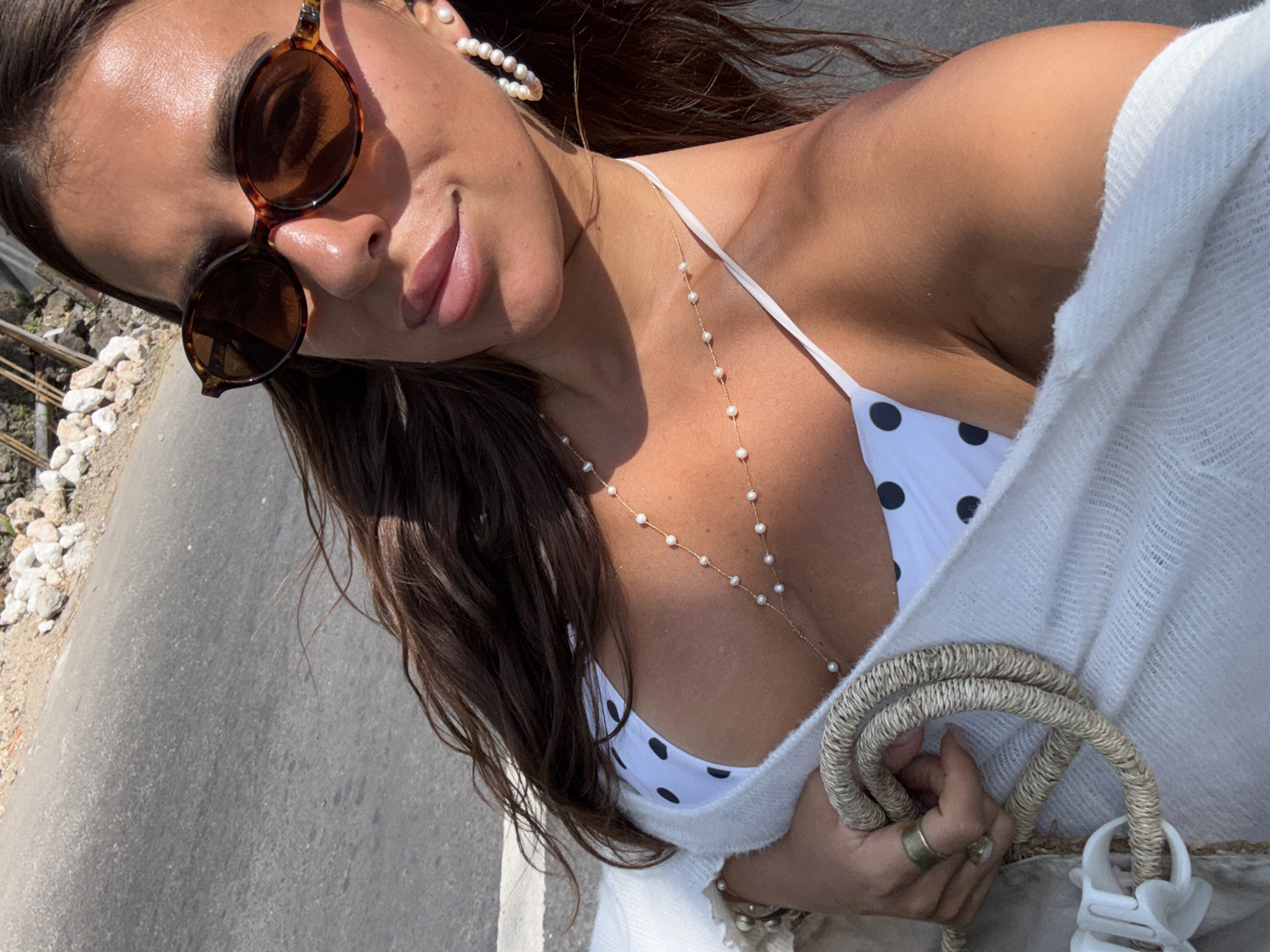 A woman with long, dark hair, wearing sunglasses and a white polka dot bikini, is captured in a close-up, selfie-style shot. She has on pearl earrings and a delicate pearl necklace. A light-colored, textured wrap is draped over her shoulder, and she holds a woven bag with rope handles. Her right hand, adorned with rings, rests on the bag. The background is a sunlit asphalt road with a rocky, uneven embankment on the right side. This scene is consistent with outdoor locations in Kuta Selatan, Indonesia, known for its beaches and roads.