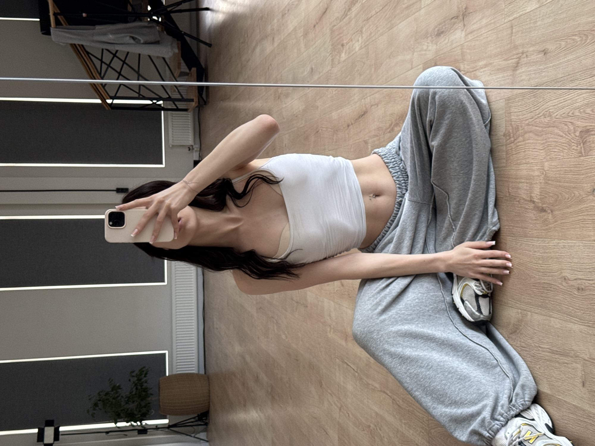 A young woman is sitting on a wooden floor in a studio space, taking a mirror selfie with her phone. She is wearing a white crop top and grey sweatpants, with athletic shoes on her feet. In the background, there is a plant in a wicker pot, a radiator, a staircase with a railing, and vertical lighting fixtures. The floor appears to be polished wood. The overall impression is of a casual, indoor environment.