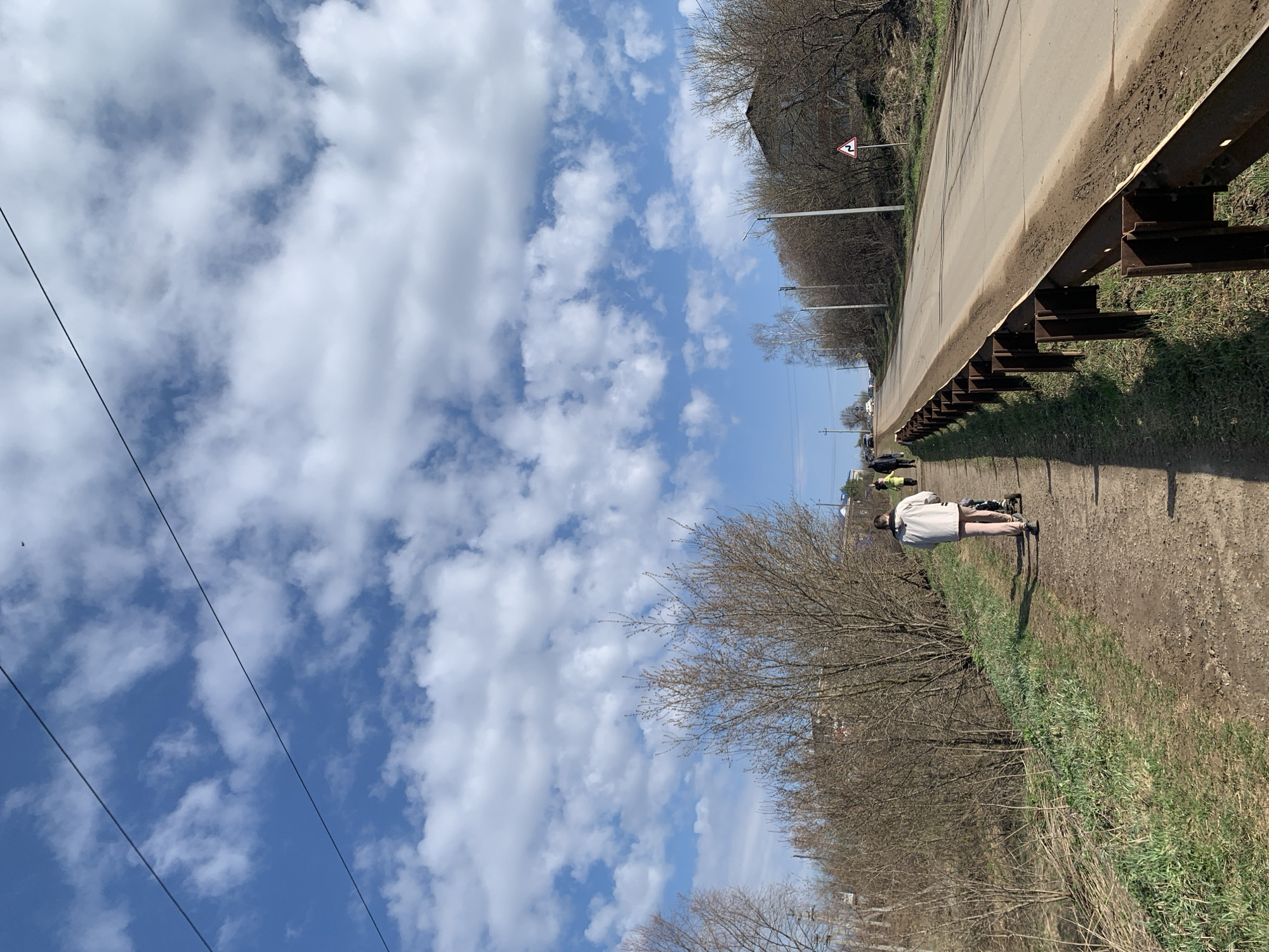 This vertical image depicts an outdoor scene under a bright, partly cloudy sky, likely in a rural or semi-rural area near Фоки, Russia. The upper portion of the frame is dominated by a clear blue sky interspersed with numerous white, puffy clouds. Power lines are visible stretching diagonally across the sky in the top left.
Running from the middle to the right side of the image is a paved road or elevated pathway, bordered by a long, rusty brown metal guardrail supported by metal posts. Below this barrier, on the left side, is a dirt path or ground covered with sparse, dry grass and leafless trees and bushes, suggesting late winter or early spring.
One person is clearly visible on the dirt path in the mid-ground, standing with their back mostly to the camera. They are wearing a light-colored jacket or coat and light-colored pants. A second person is faintly discernible further down the path.
Notable details include a triangular road sign with a red border and white background, displaying a black symbol of a winding road. There appears to be a faint number "2" at the bottom right of this sign. Utility poles and possibly streetlights line the road further into the distance, and several vehicles are vaguely visible further along the road. The scene is illuminated by bright daylight, with no discernible emotional tone.