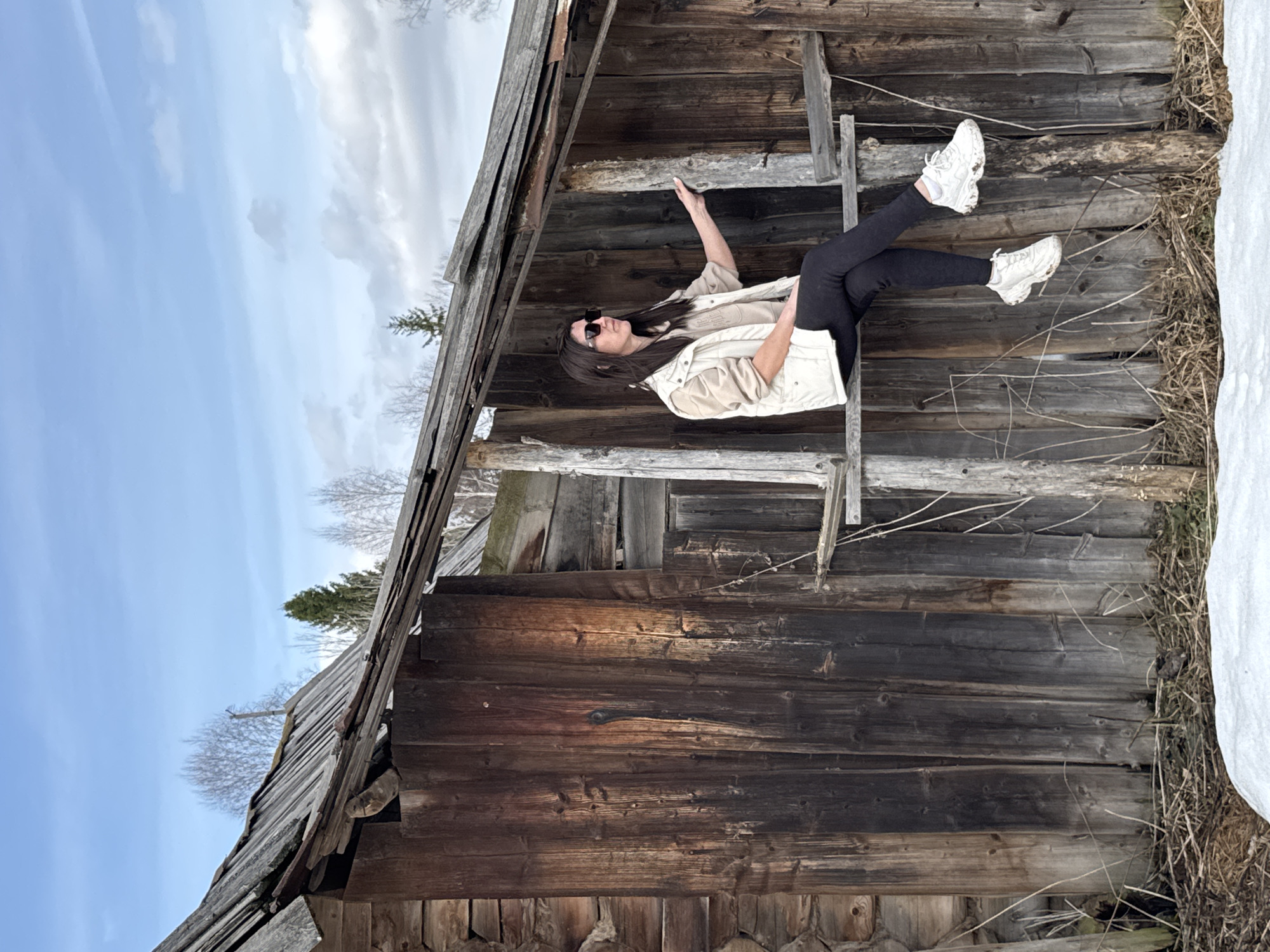 The photograph, taken in Вассята, Russia, shows a woman positioned within the decaying structure of a wooden building. She is wearing sunglasses, a tan shirt, a white vest, black pants, and white sneakers. The woman is lying on a support beam in a horizontal manner, with one arm raised. The building appears dilapidated with exposed wooden beams and incomplete walls. Patches of snow are visible outside the structure. The sky is blue with wispy clouds. The image is rotated ninety degrees clockwise.