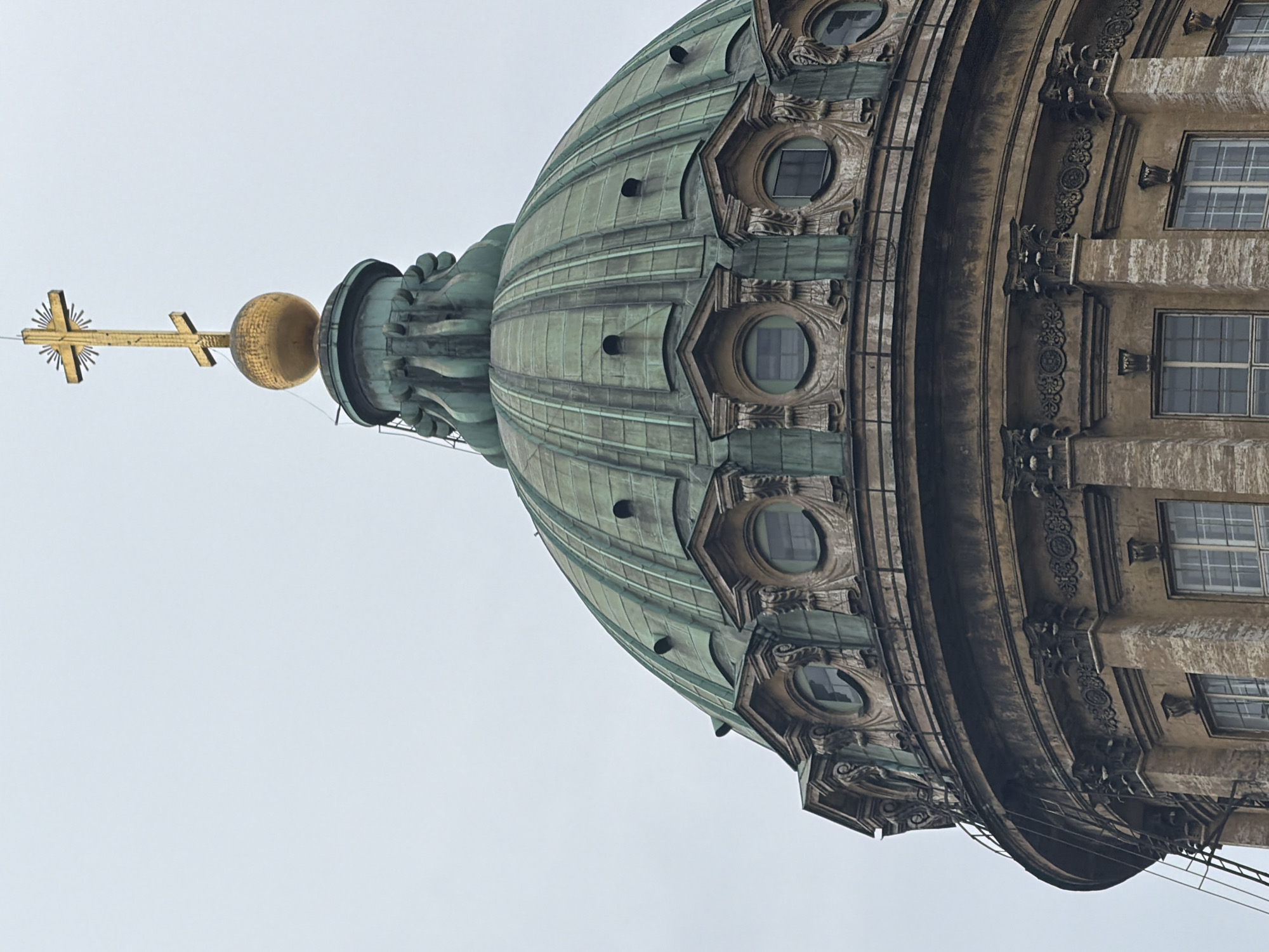 The image depicts the upper dome and architectural elements of the Kazan Cathedral, located in Saint Petersburg, Russia. The primary subject is a large, patinated green, ribbed hemispherical dome. Directly below the dome's base, a band of light-colored stone is visible, featuring multiple ornate round dormer windows (oculi) with dark, grid-patterned panes, framed by intricate carved stonework. Above the main dome, a narrower, fluted dark green cupola supports a large, gilded spherical orb. Mounted atop this orb is a prominent gilded Russian Orthodox cross, distinguished by a third slanted bottom bar and radiating spikes from its center. The visible lower section of the cathedral's facade, beneath the dome, is constructed from light-colored, weathered stone exhibiting striations and dark staining. This section incorporates classical architectural details, including engaged columns with Corinthian capitals, decorative friezes with circular motifs, and rectangular multi-paned windows. A dark metal ladder or scaffolding structure is partially visible ascending the curved facade on the lower-left side. The sky above is uniformly light grey, indicating an overcast condition. No human subjects or active events are present in the scene.