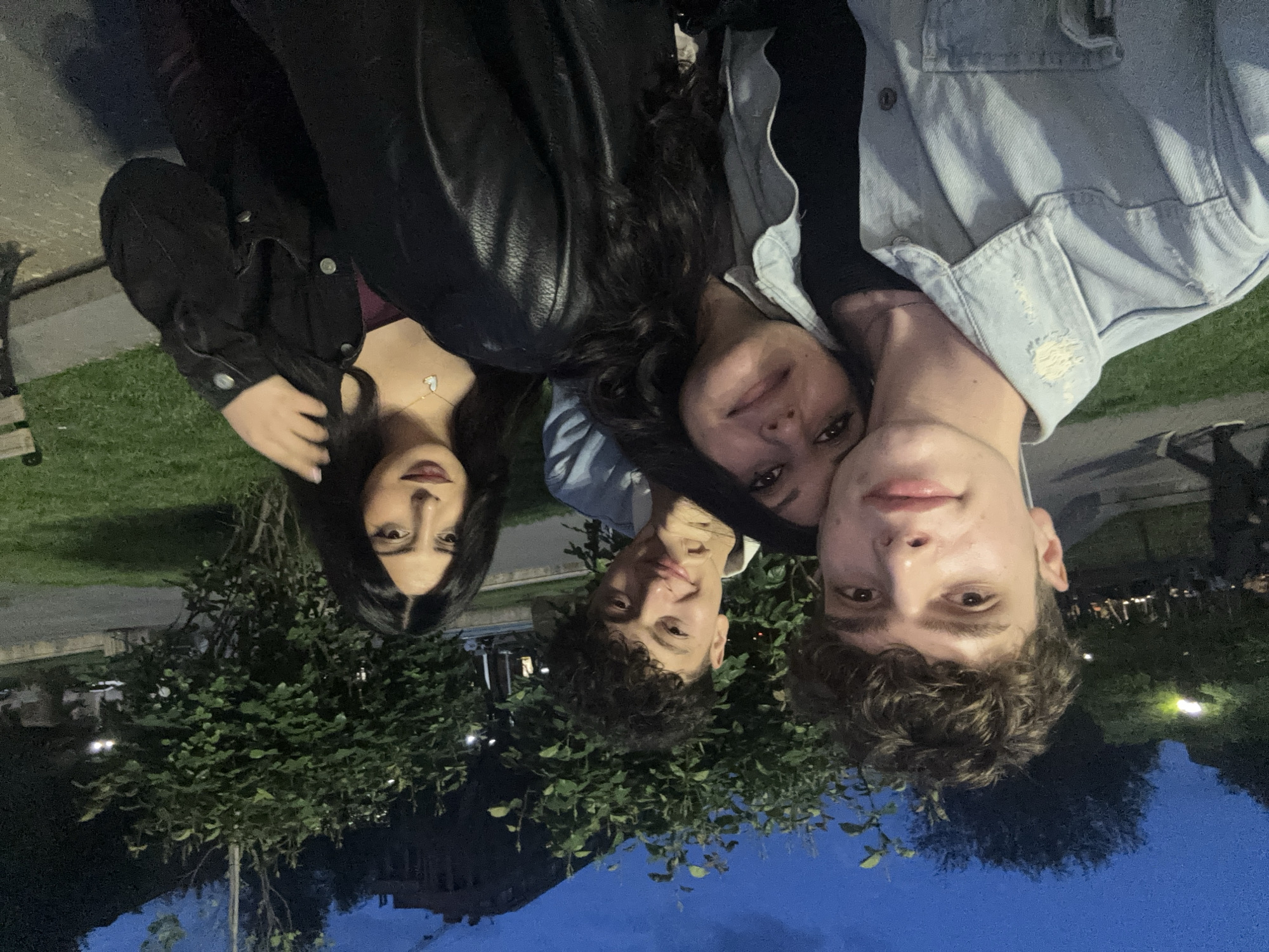 A group of four young people is posing for a photograph outdoors at night. The person on the left has long, dark hair and is wearing a black jacket. Next to her, another person is wearing a black leather jacket. To their right, a person with curly dark hair smiles. Finally, a young man wearing a light blue denim jacket is on the right. They are standing in front of a small, leafy tree or bush, with a city street and buildings visible in the background under a twilight sky. Based on the visible architecture, including buildings and streetlights, and the style of vegetation, it is likely that this picture was taken in Yerevan, Armenia.