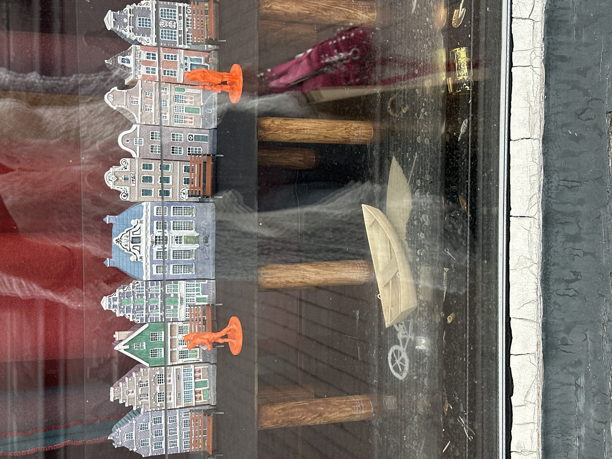 The image depicts a miniature street scene, likely a diorama, displayed behind glass. A row of colorful, Dutch-style houses with detailed facades lines the top of the scene. In front of the houses, there are two orange figurines, one appearing to be an adult holding a child. Miniature park benches are placed in front of some of the houses. Below this upper level, the scene transitions to a darker, more subdued area with wooden posts, suggestive of a pier or bridge supports. A small wooden boat rests on what appears to be water or a reflective surface in the lower portion of the image, along with a miniature bicycle. The overall impression is a stylized representation of an Amsterdam canal scene. The background visible through the glass is blurred, but appears to be fabric with a reddish hue. The foreground, below the display, shows weathered, cracked paint on a ledge.