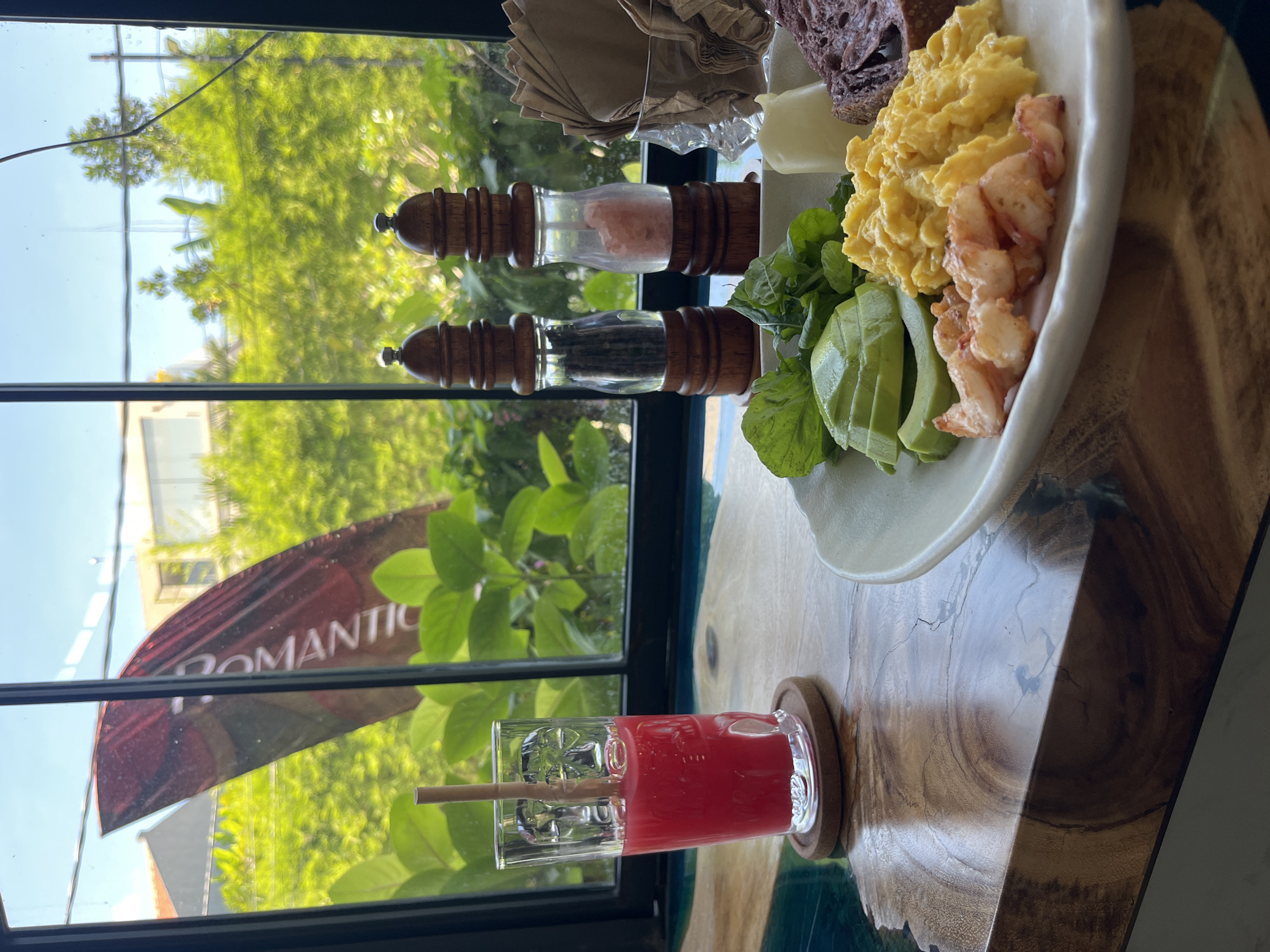 Captured from a high-angle perspective, the scene features a breakfast setup adjacent to a window with a partial view of the outdoors. The breakfast includes a plate with scrambled eggs, lobster pieces, avocado slices, and bread. Adjacent to the plate are salt and pepper shakers. A beverage in a glass is placed on a coaster. The outdoor scenery visible through the window includes trees and building. A flag with the word "Romantic" written on it is outside of the window. The photograph was taken in North Kuta, Indonesia.