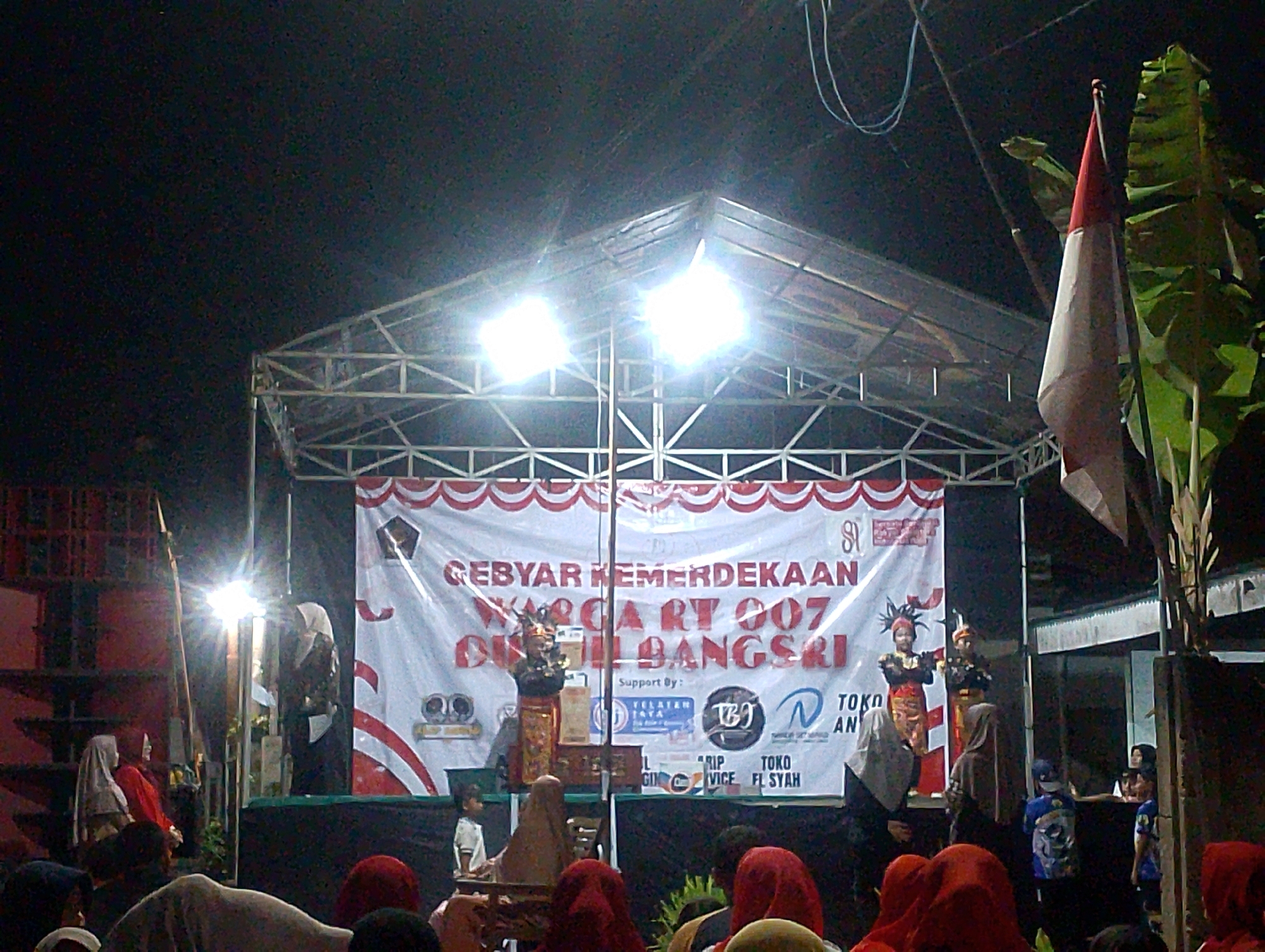 This is a nighttime outdoor photograph of a stage setup for what appears to be a community event. A large white banner with red and white trim stretches across the back of the stage, displaying text that reads "GEBYAR KEMERDEKAAN WARGA RT 007 DI NGURUS BANGSRI". Below this text, there are several logos and sponsorship information. Two individuals in traditional attire, possibly performers, stand on the stage. A small child in a white shirt is also visible on the stage. In the foreground, a crowd of people, many wearing headscarves, are gathered, looking towards the stage. A tall pole with a flag, likely the Indonesian flag due to its red and white colors, is positioned to the right of the stage. Lush green foliage from a banana tree is visible on the far right side of the frame. The stage itself is a metal structure with a translucent roof, illuminated by bright lights. The scene suggests a lively local celebration, possibly in Plumbungan, Indonesia, given the context provided.