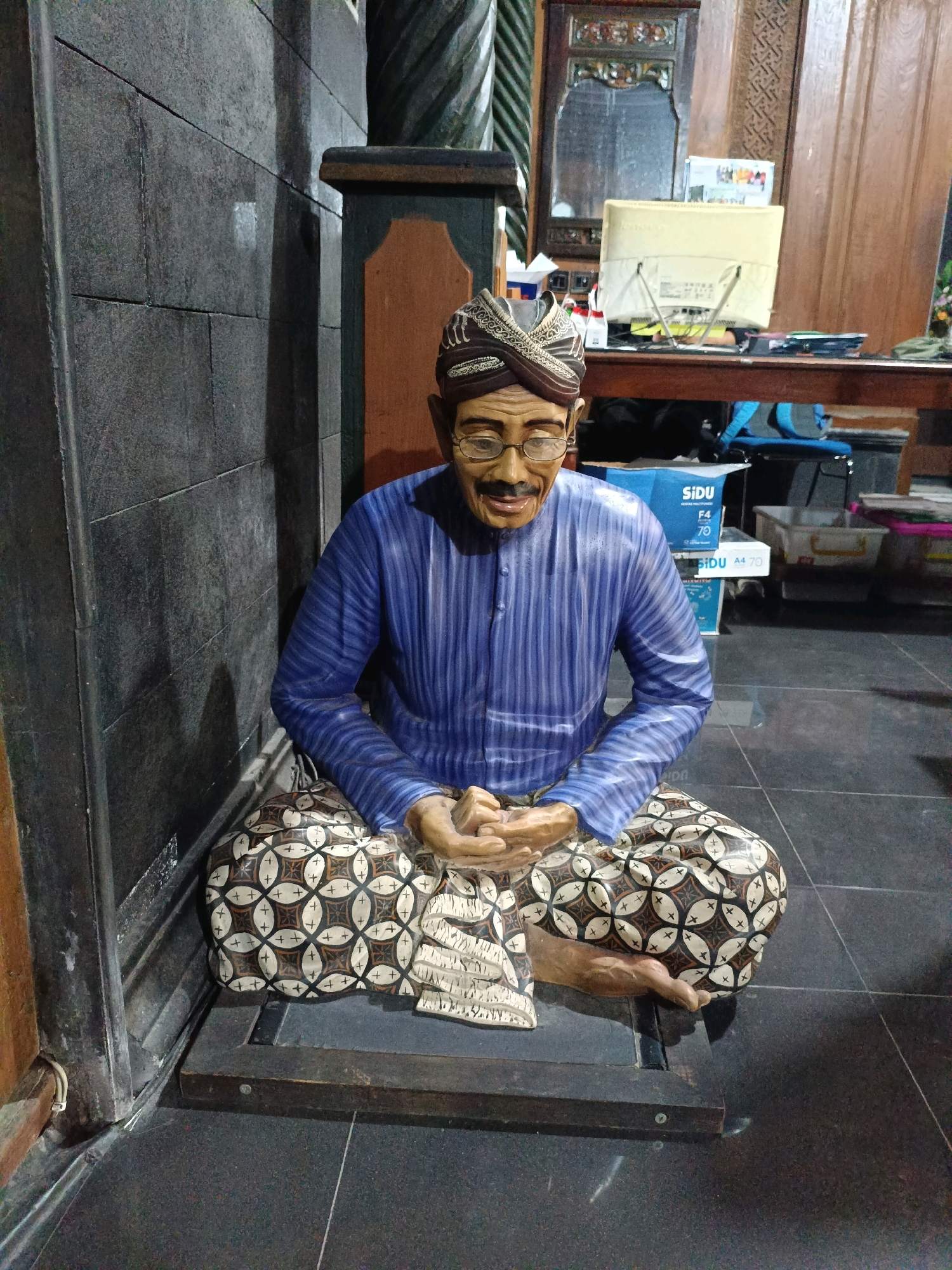 A full shot captures a realistic, life-sized statue of a man sitting cross-legged on a dark, tiled floor in what appears to be an indoor setting. The man is dressed in traditional Indonesian attire: a blue striped long-sleeved shirt, a patterned sarong with a circular motif in shades of brown and cream, and a decorative head covering. He wears glasses and has a mustache and goatee. His hands are clasped in his lap. The statue is positioned on a dark wooden platform. In the background, a dark stone wall is visible on the left, and further back, a wooden desk with a computer monitor and papers is situated against a wooden paneled wall. The lighting in the scene is dim, creating shadows. This scene is located in Purwomartani, Indonesia.