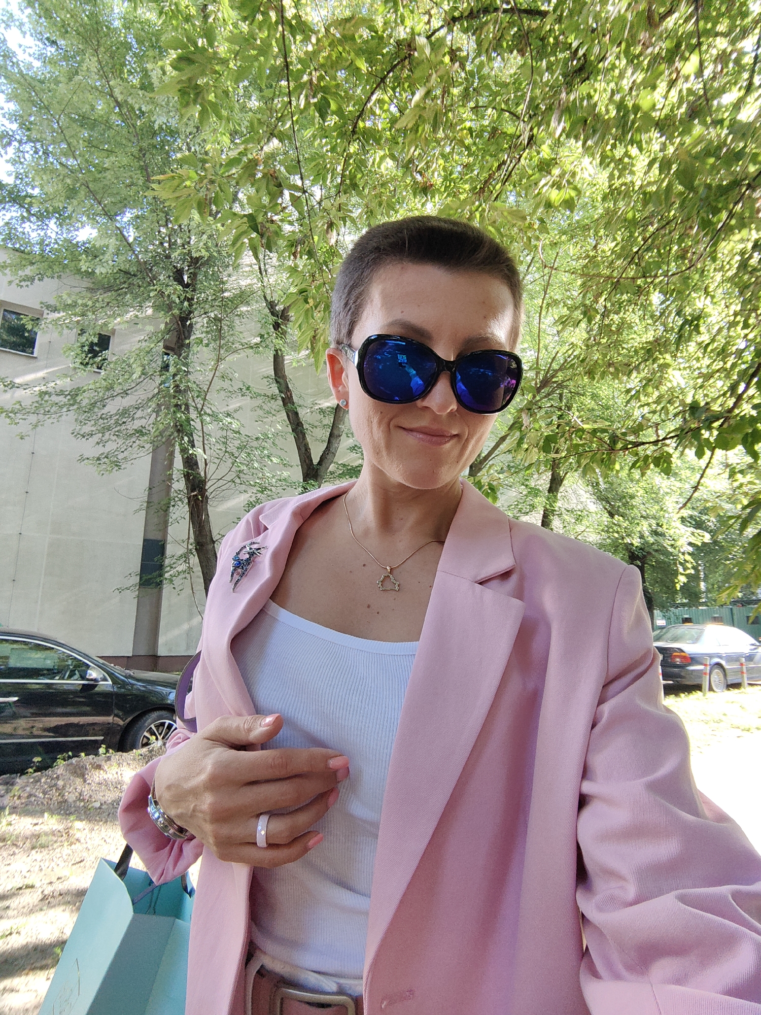 A woman with short hair and large, dark sunglasses stands outdoors in front of a light-colored building and trees. She is wearing a light pink blazer over a white ribbed tank top. A gold necklace with a geometric pendant is visible. A silver brooch with blue stones is pinned to the left lapel of her blazer. She is wearing a silver watch on her left wrist and a light-colored ring on her right ring finger. Her right hand is resting on her chest. She is holding a light blue shopping bag in her left hand. A dark-colored car is parked to the left of the frame, and another dark-colored car is visible in the background to the right. The environment appears to be a street or parking area in a city.