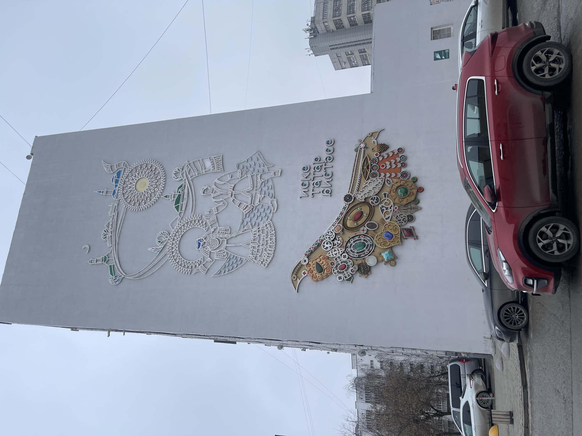 The image shows a building with a large mural on its facade. The mural features stylized, decorative depictions of two birds or mythical creatures, along with text in a Cyrillic script. The left-hand creature is primarily white, while the right-hand one incorporates gold and various colored decorative elements resembling jewels. The building is located in Kazan, Russia. Parked along the side of the building, partially obscuring the mural, are several cars, including a red SUV, a dark-colored sedan, and a white vehicle. The sky is overcast. The street is wet.