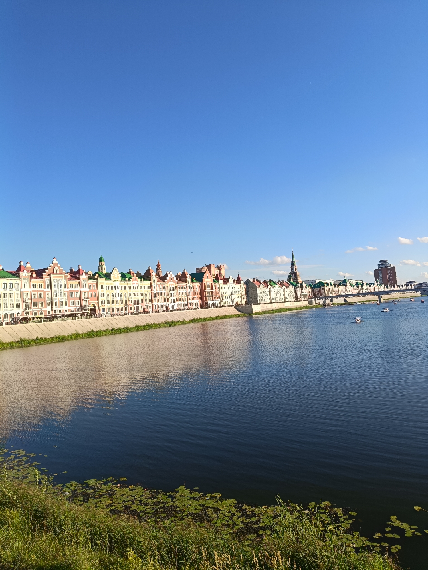 This is a wide-angle, eye-level, outdoor, daytime photograph of the embankment in Yoshkar-Ola, Russia. The scene features a river with boats, a promenade, and colorful buildings along the waterfront. The sky is a clear, vibrant blue.
In the foreground, the river occupies the lower half of the image, with a gentle ripple pattern on its surface. Lily pads and tall green grass are visible along the near bank. The river reflects the sky and the buildings.
The middle ground showcases a concrete embankment or quay wall, separating the river from the promenade. Along the promenade, an array of buildings with distinctive architectural styles stretches across the frame. These buildings are painted in various shades of pink, yellow, white, and red, with green roofs and decorative elements. Some buildings have multiple stories, with windows and balconies. Several outdoor seating areas, possibly for cafes or restaurants, are visible along the promenade. A tall, slender green spire rises from one of the buildings, resembling a church tower. Further down the embankment, a modern multi-story building with a reddish-brown facade and a dark-topped structure is visible. A bridge spans the river in the distance. Small boats, including what appear to be pedal boats, are scattered across the river.
The background consists of the expansive blue sky, with a few wispy white clouds. A single bird is visible flying high in the sky. The overall impression is a picturesque urban waterfront on a sunny day.