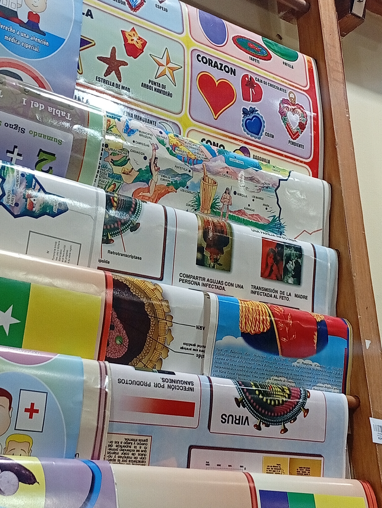 The image shows a close-up, eye-level, indoor shot of a wooden shelf unit displaying several rolled-up educational posters. The posters are layered on top of each other, with the top one partially obscuring the ones below. The posters feature colorful illustrations and text in Spanish, covering topics such as marine life (starfish, Christmas tree ornament), geometric shapes (heart, circle, oval), a map of Venezuela with place names and illustrations, and medical information about diseases like AIDS and viruses. One poster, likely about the history of Venezuela, mentions "Juan Griego" and other locations. The wooden shelves are visible on the right side of the frame, and the background is out of focus, suggesting an indoor setting, possibly a school or a shop. The lighting appears to be from an artificial source, creating a bright and evenly illuminated scene.