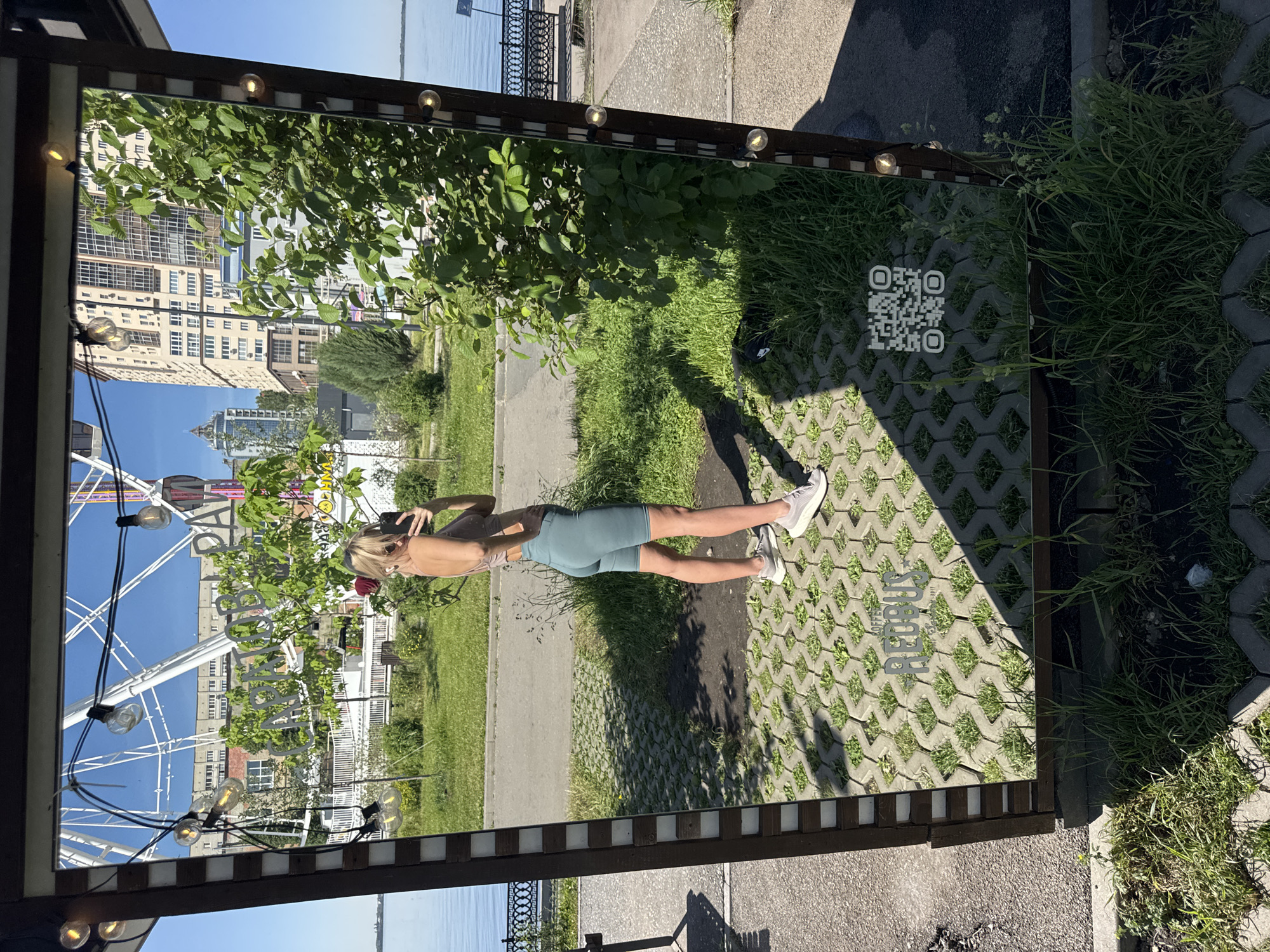 The image shows a woman standing in front of a large mirror, taking a picture of herself with her phone. The mirror reflects a view of Saratov, Russia, including a ferris wheel, buildings, and greenery. The woman is wearing athletic attire: a crop top and shorts. The ground in front of the mirror is paved with interlocking concrete blocks with grass growing in the gaps. There is a QR code on the paving near the mirror. The mirror has a wooden frame with light bulbs around it. The background consists of a sidewalk and grass.