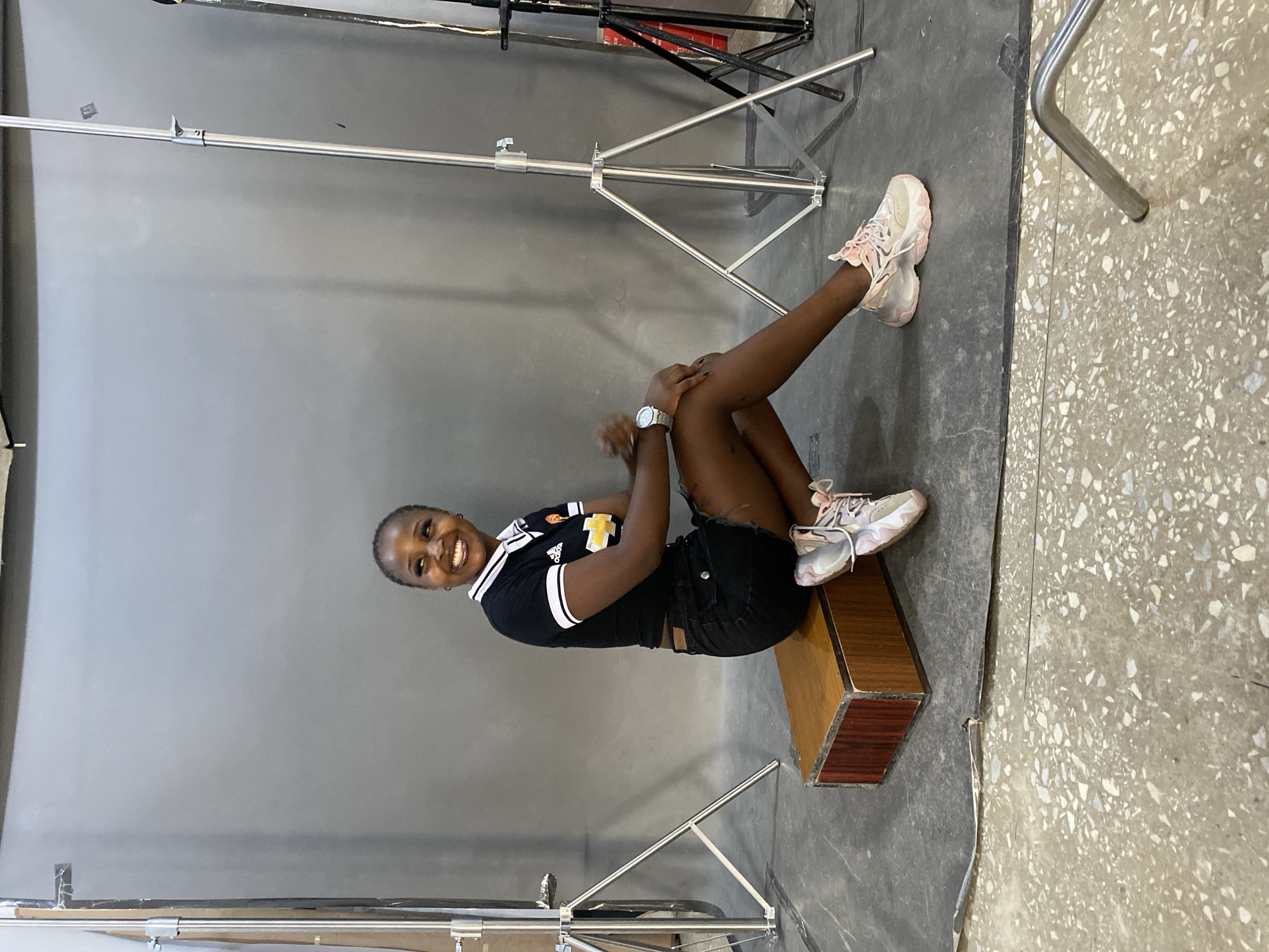In a studio setting with a grey backdrop, a young Black woman is seated on a wooden crate. She is wearing a black t-shirt with a white collar and trim, black denim shorts, and pink and white athletic shoes. Her hair is pulled back, and she is smiling, looking to her left. The studio floor is a textured grey mat with a speckled concrete floor visible in the foreground. Two lighting stands are positioned behind her, one on each side. The scene appears to be a casual portrait shoot. The location in Gura Top, Nigeria, is not discernible from the visual information provided in the image.