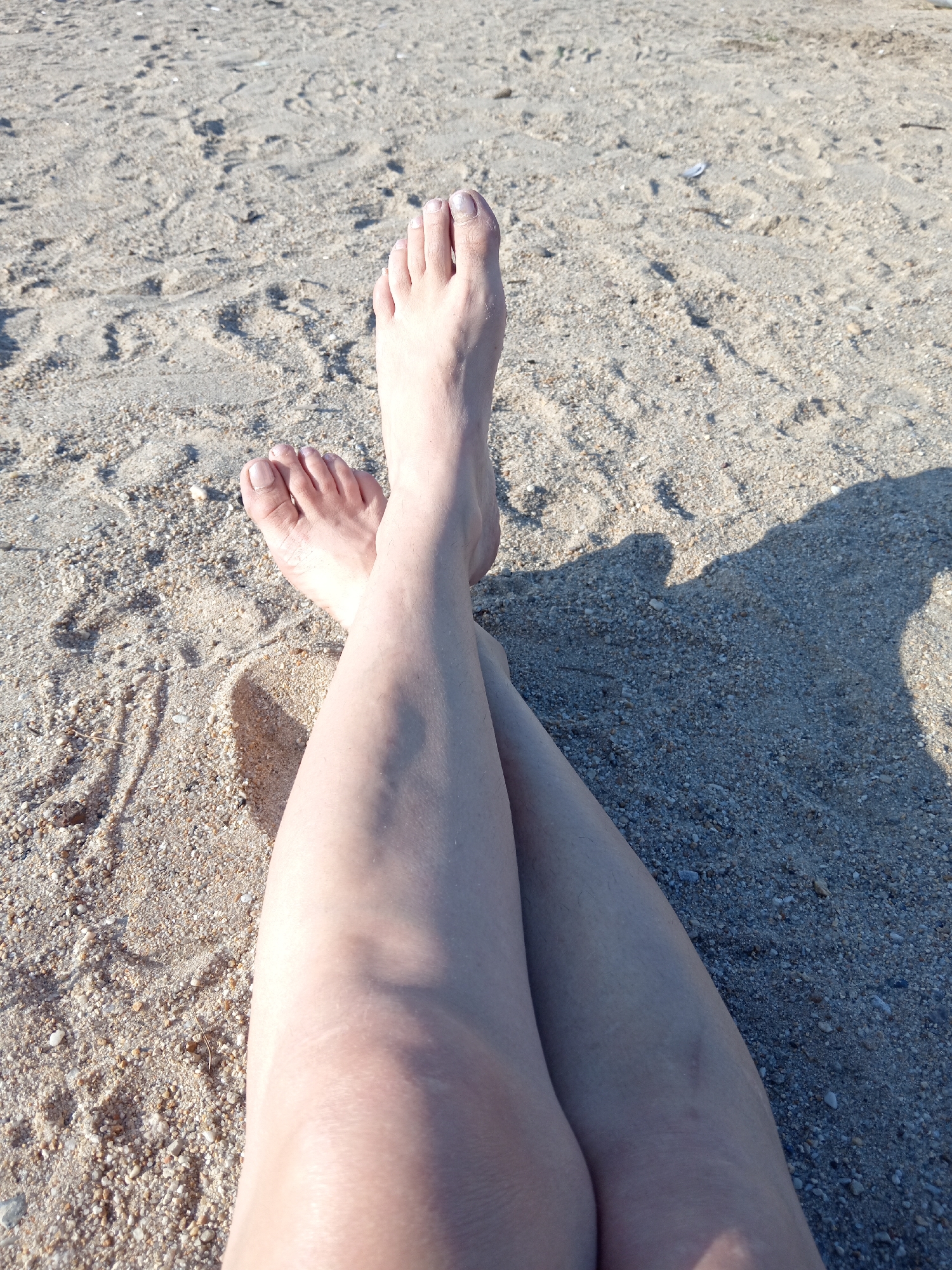 A close-up aerial shot shows a pair of legs crossed at the ankles, resting on a sandy beach. The skin is lightly tanned, and fine hairs are visible on the legs. The toes of both feet are visible, with the nails appearing unpolished. The sand is a light brown color with small pebbles and shell fragments mixed in. A shadow, cast by an unseen object or person, is visible on the sand to the right of the legs. The scene is set on a beach in Belogorsk, Russia.
