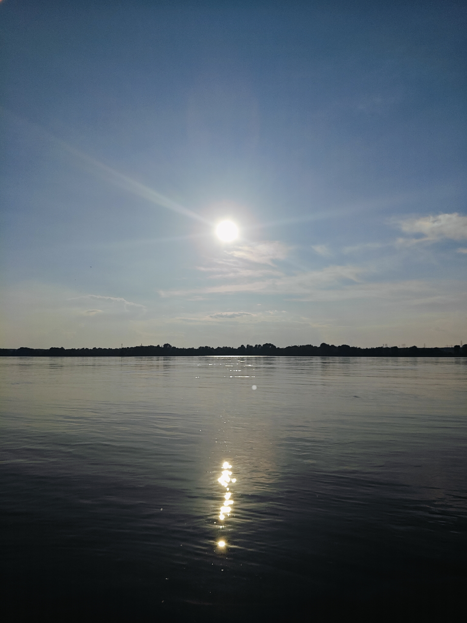 The image depicts a wide, calm river with a distant, dark treeline on the horizon under a bright, clear blue sky. The sun, a bright white orb with rays extending outwards, is positioned in the upper central portion of the frame. A shimmering reflection of the sun stretches across the water's surface, creating a path of light. The water appears still, with subtle ripples. The treeline is silhouetted against the lighter sky, suggesting it is either dawn or dusk. The scene appears to be a natural landscape. It is highly probable this image was taken in or around Chaikovsky, Russia, known for its location on the Kama River, which matches the depiction of a wide river. There are no people or discernible objects in the water or on the distant shore apart from vegetation and what appear to be distant power lines.