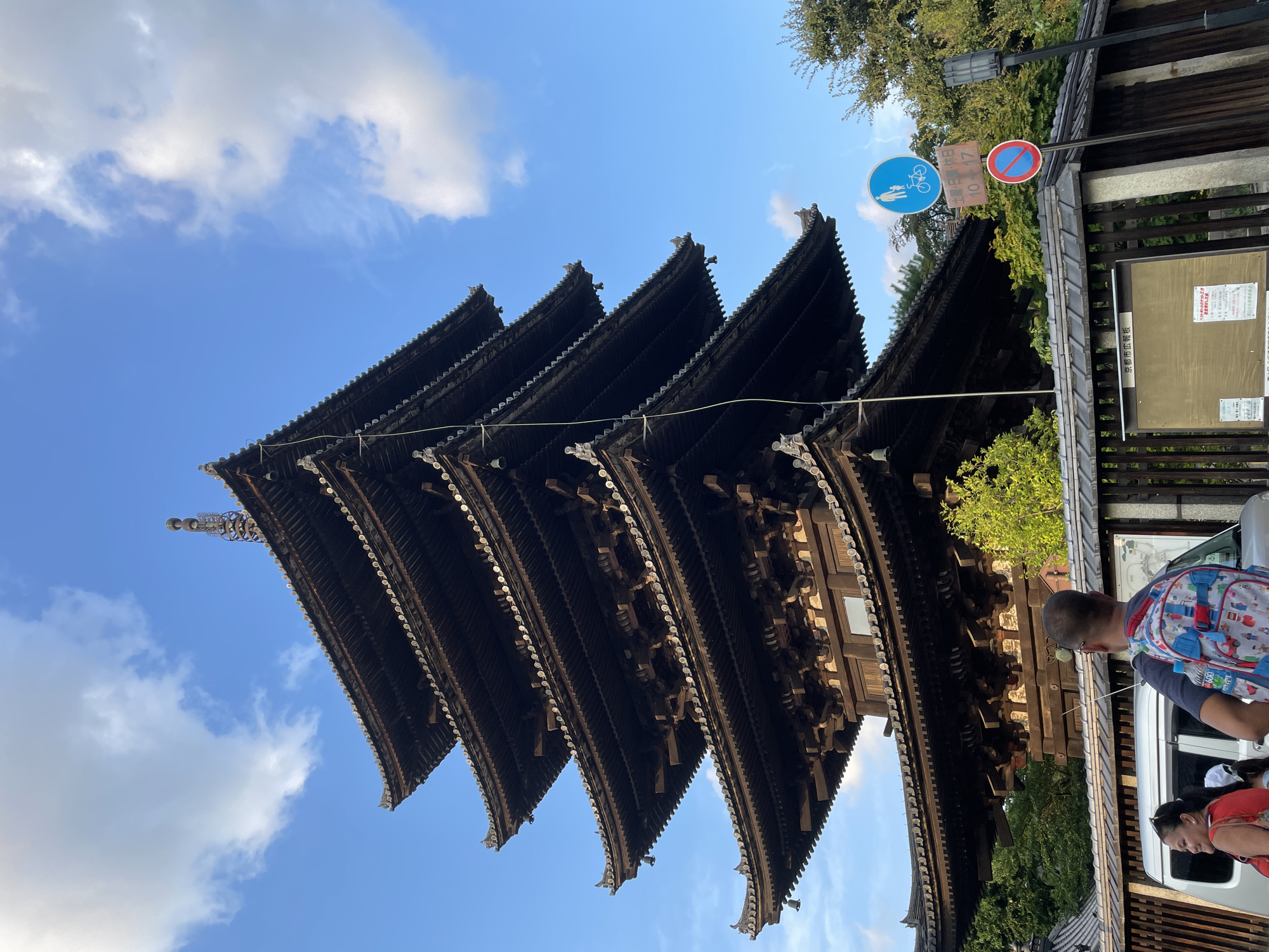 The image shows the To-ji Temple's five-story pagoda in Kyoto, Japan, under a partly cloudy blue sky. The wooden pagoda, a significant landmark in Kyoto, features multiple tiered roofs with intricate detailing and small bells hanging from the eaves. In the foreground, a wooden fence encloses a bulletin board. To the right of the fence, traffic signs are visible, including a blue circular sign with a pedestrian and bicycle symbol, and a red circular sign with a diagonal bar, indicating no parking. Two people are partially visible on the left side of the image, near a silver vehicle. One person, a woman wearing a red top, is looking to the left. The second person, a man wearing a backpack, is facing away from the camera and looking towards the pagoda. The environment suggests a public area with vegetation and trees surrounding the temple grounds.