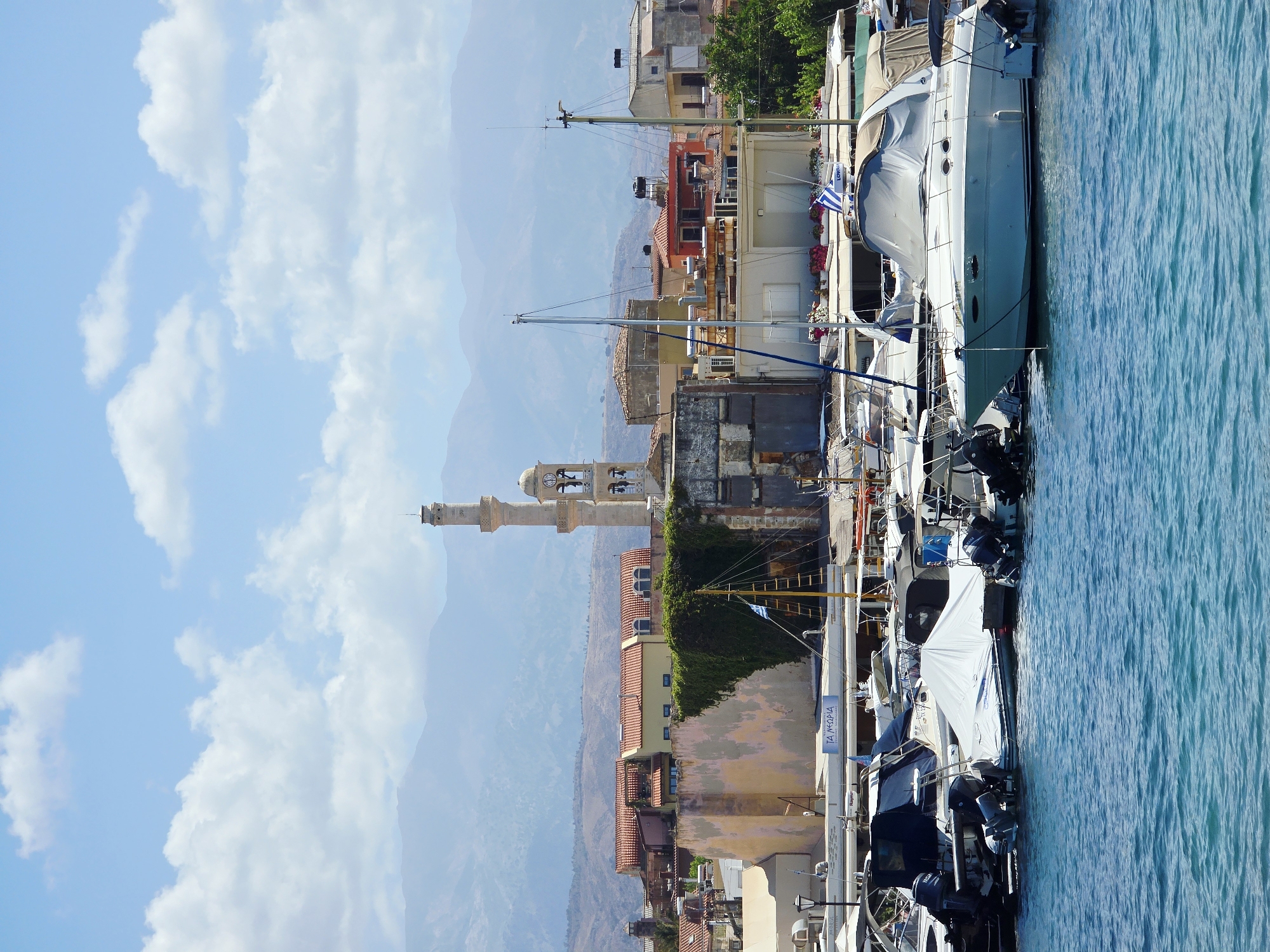 The image shows a harbor in the city of Chania, Greece, with a body of water in the foreground and boats docked along a quay. In the background, buildings with terracotta roofs are visible, along with a prominent minaret-like structure with a clock face and bell tower, which is part of the historical Grand Arsenal. Mountains are visible in the distant background, softened by atmospheric haze. The water is a vibrant turquoise color, and several boats of various sizes are moored. Some boats are covered, while others are uncovered, revealing their decks and equipment. The sky is blue with scattered white clouds. The overall scene depicts a sunny day in a coastal European city with a rich history.