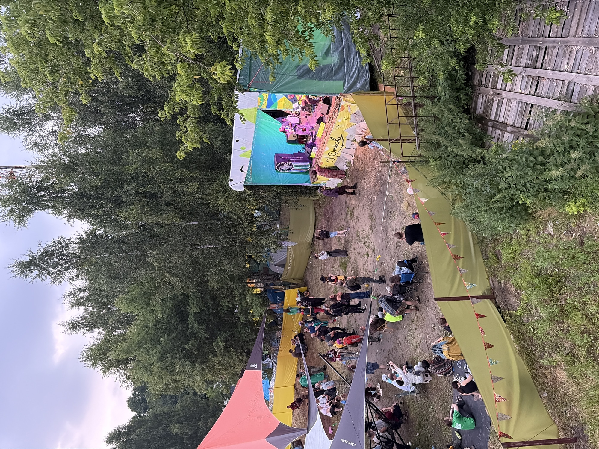 The image depicts an outdoor music festival in Il'inskiy Pogost, Russia. A stage with a band performing is the central focus, set against a backdrop of trees and tents. A crowd of people is gathered in front of the stage, some sitting on the ground, others standing, and a few seated in chairs. Various tents and temporary structures are visible throughout the scene, suggesting a larger festival area. The ground is dirt and grass, with some wooden structures and railings. The overall atmosphere appears to be relaxed and casual, with people enjoying the music and the outdoor setting. The lighting suggests it is late afternoon or early evening.