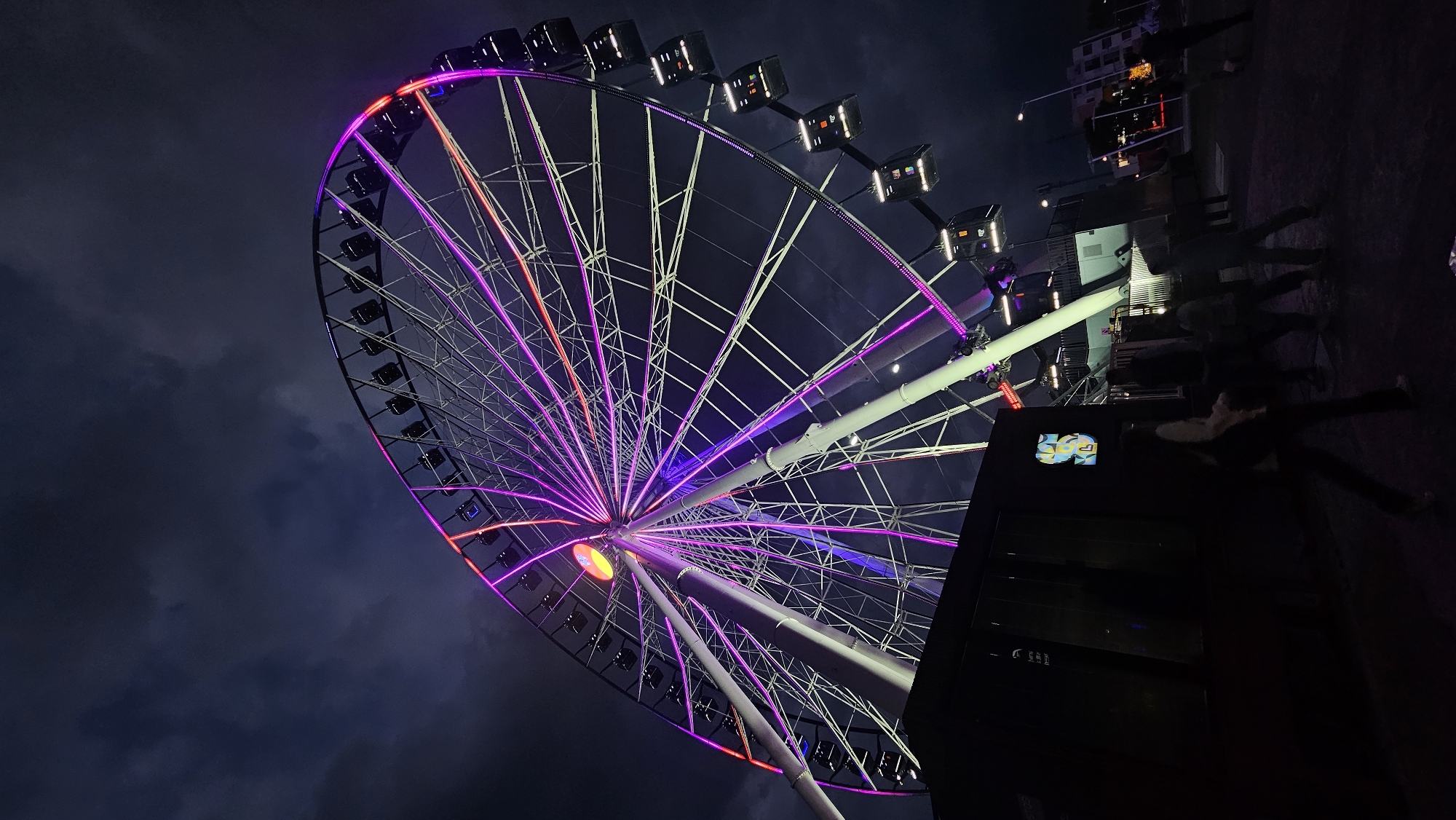 The image depicts a Ferris wheel at night, presumably located in Puebla, Mexico. The Ferris wheel is illuminated with a variety of colors, predominantly purple, pink, and orange. Its steel frame is visible against the dark sky, and each of the enclosed passenger cabins is lit from within. The base of the Ferris wheel is partially obscured by a dark structure, possibly a building, with a sign visible on its side. Some people are walking in the lower right corner of the frame; they appear to be in motion, causing motion blur. The sky is a dark, overcast blue, suggesting it is nighttime.