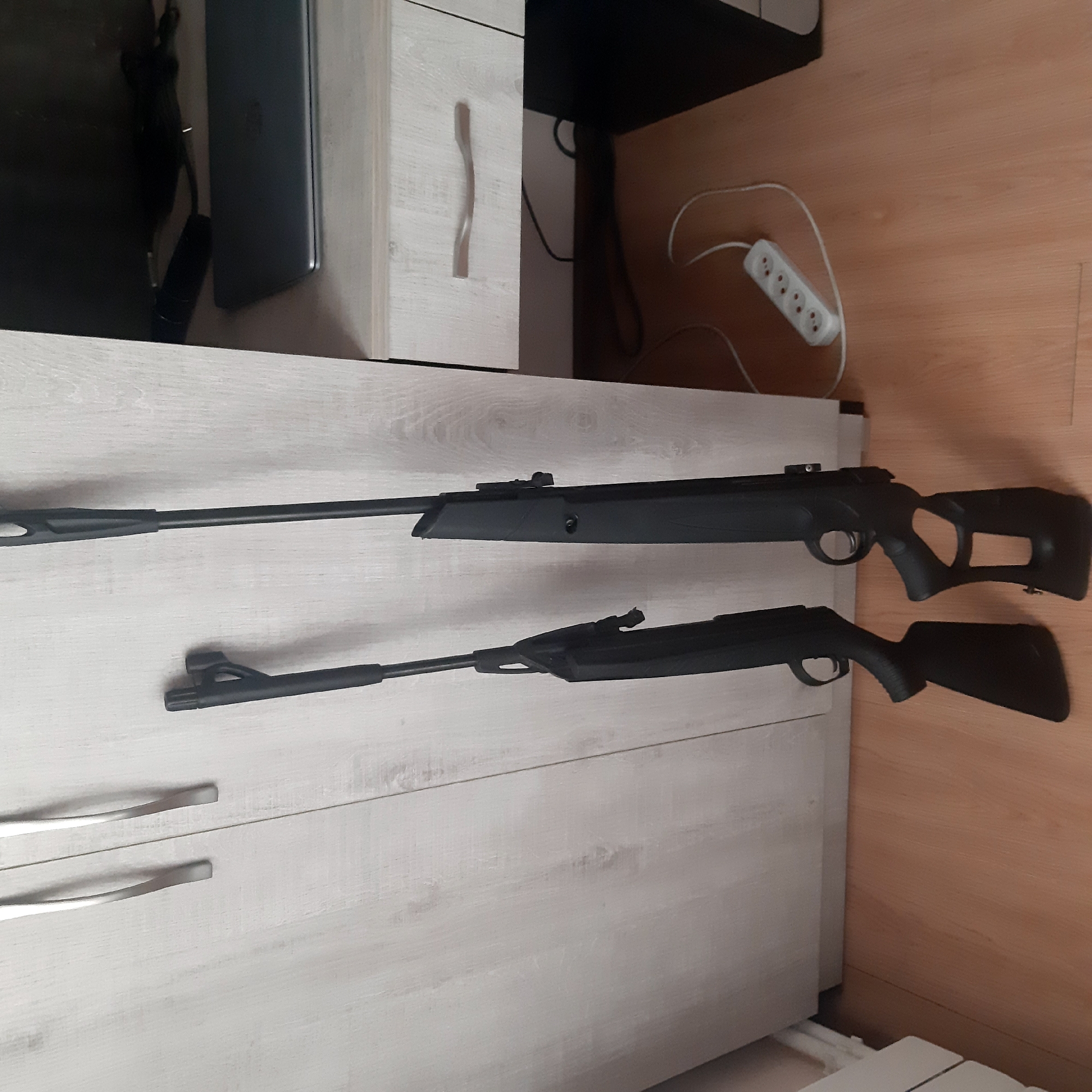 Two black air rifles are lying horizontally on a light-colored wooden surface, possibly a dresser or table. An electrical power strip is visible in the background to the right, on a wooden floor. The scene is indoors, and no people are visible. There is no information to suggest this image was taken in Yeghegnadzor, Armenia.