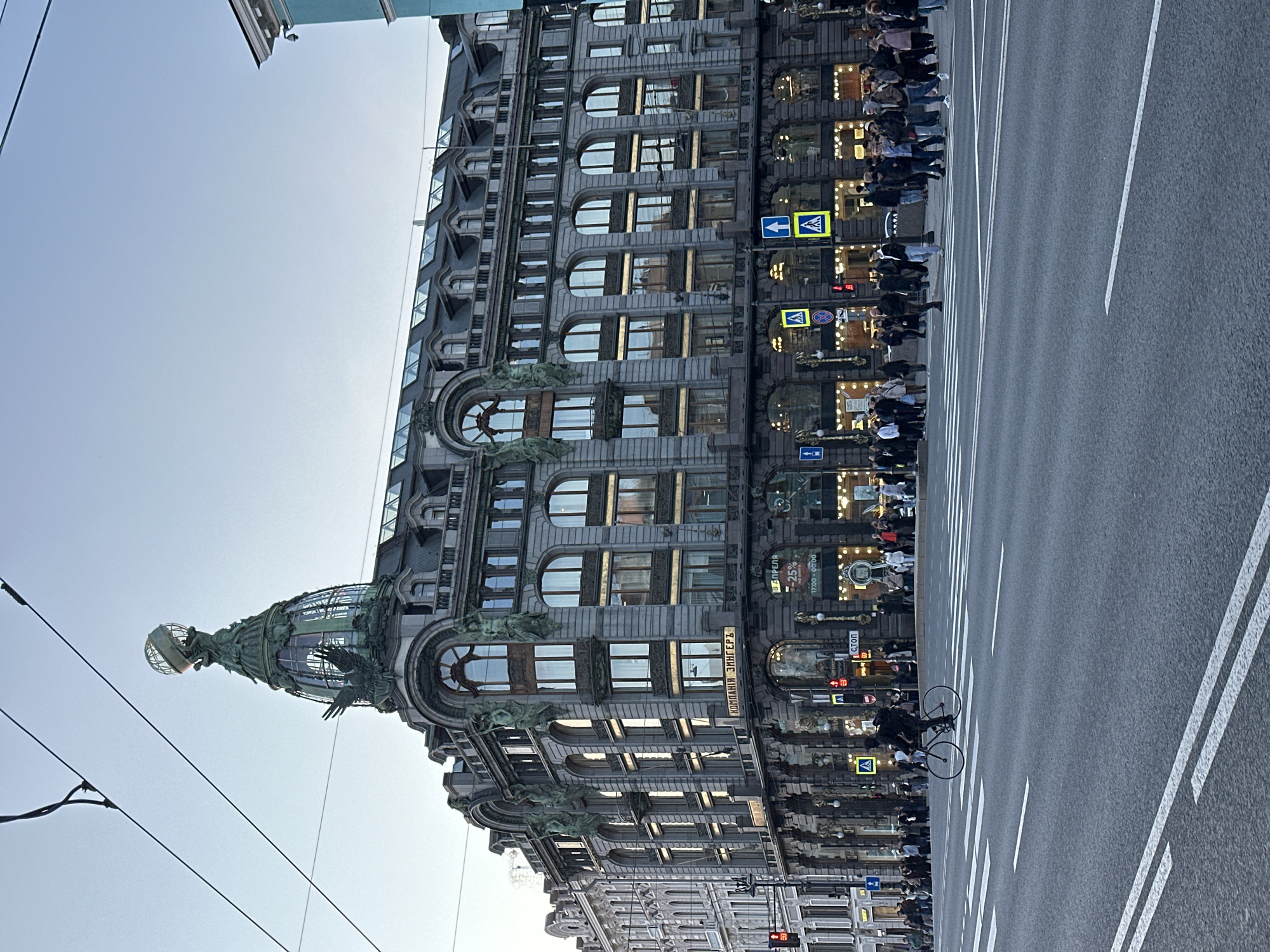 The image showcases the Singer House in Saint Petersburg, Russia, viewed from a low-angle perspective. The architectural details of the building are prominent, including its ornate facade, numerous arched windows, and the globe-topped tower. The building's name "КОМПАНІЯ ЗИНГЕРЪ" is visible on the facade.
A street runs alongside the building, featuring white lane markings and a cyclist in the middle of the road. Pedestrians are walking along the sidewalk beside the building, and signs with pedestrian crossing symbols are visible.
The sky is clear and light. Overhead, electrical or tram wires are strung across the sky.