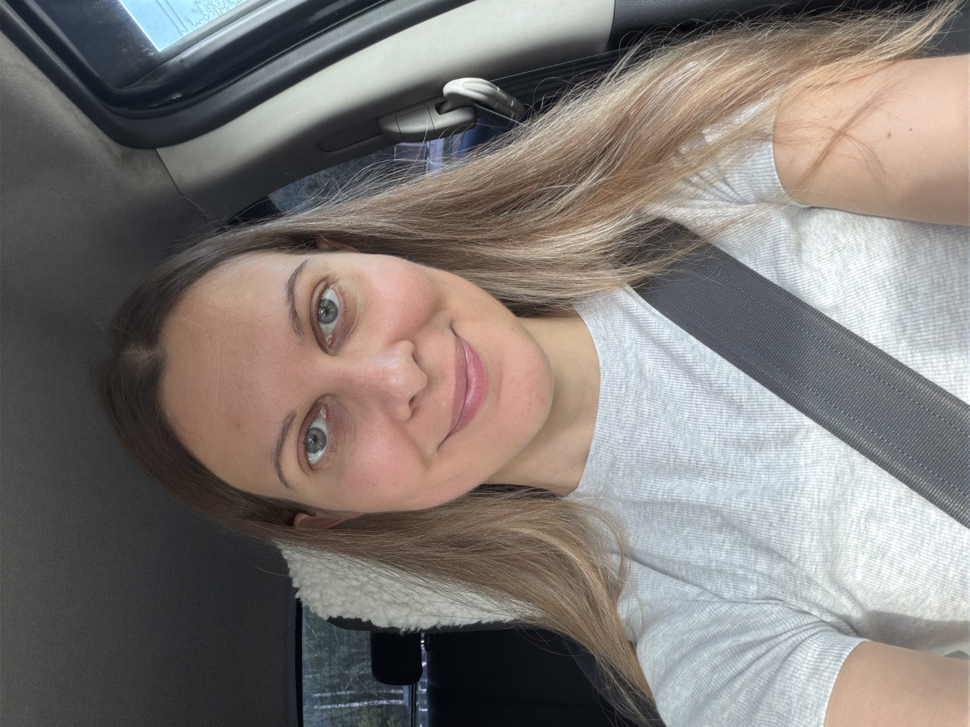 The image shows a close-up, front-facing selfie of a woman with long, blonde hair sitting in the driver's seat of a car. She is wearing a grey, ribbed t-shirt and a black seatbelt is visible across her chest. Her eyes are open, and she is smiling slightly. The background of the image is blurred, showing the interior of the car, including the dashboard and a window with trees visible outside. The context of Izhevsk, Russia, does not provide specific environmental clues to the scene.